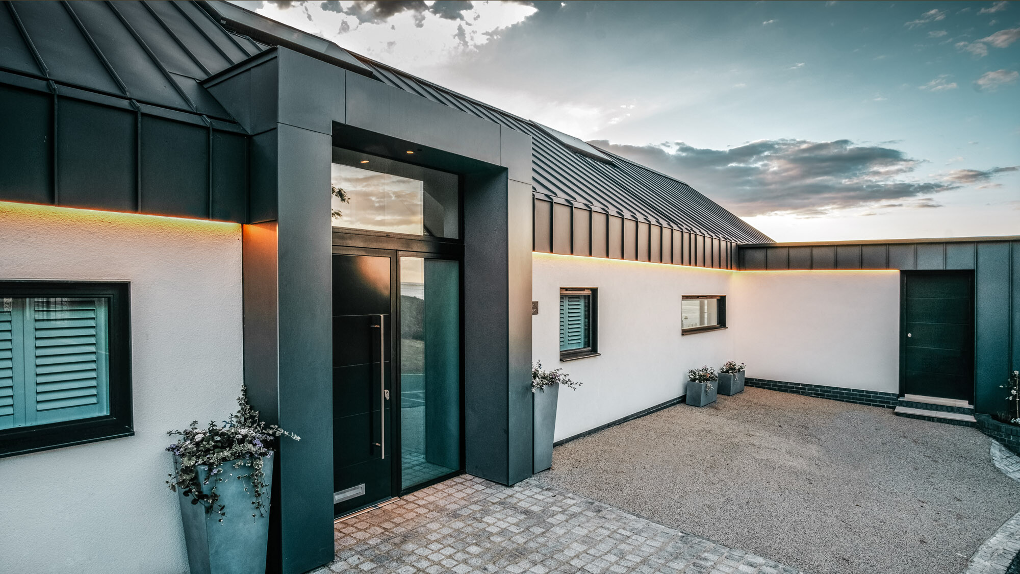 The entrance of the domicile with the dark grey aluminium, which runs seamlessly over the roof and façade and is only interrupted by the white plastered façade.