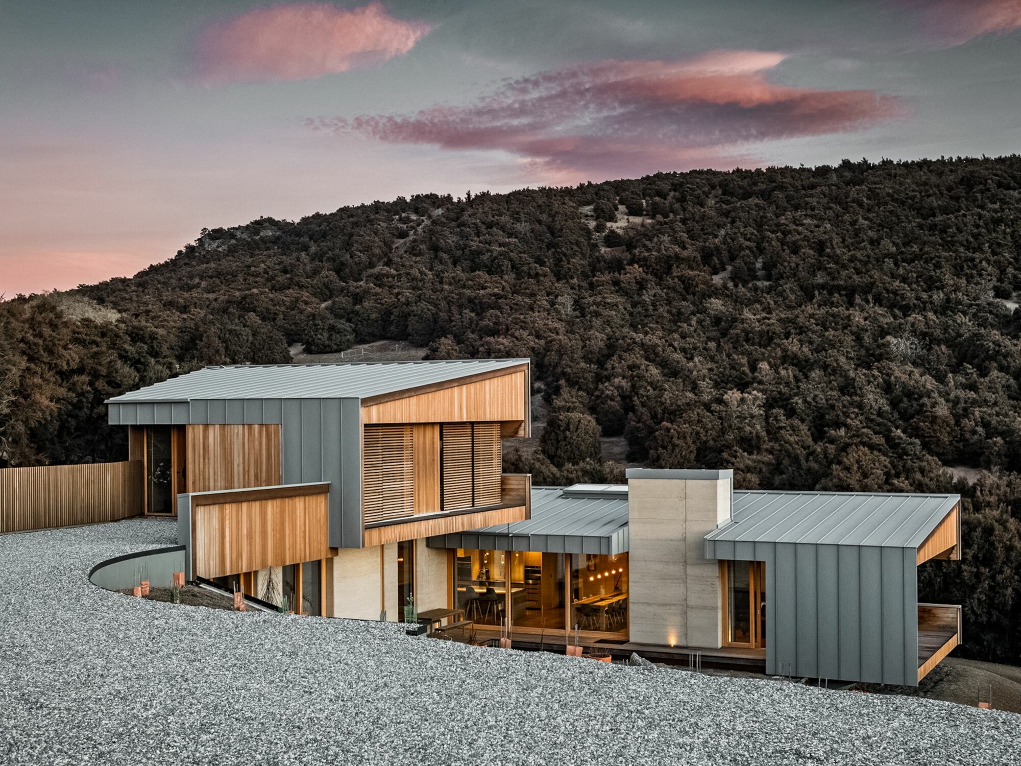 The detached house, illuminated from inside, with its driveway from a normal perspective: The alpine-looking bush landscape can be seen behind it and the sky in pastel pink and blue above it.