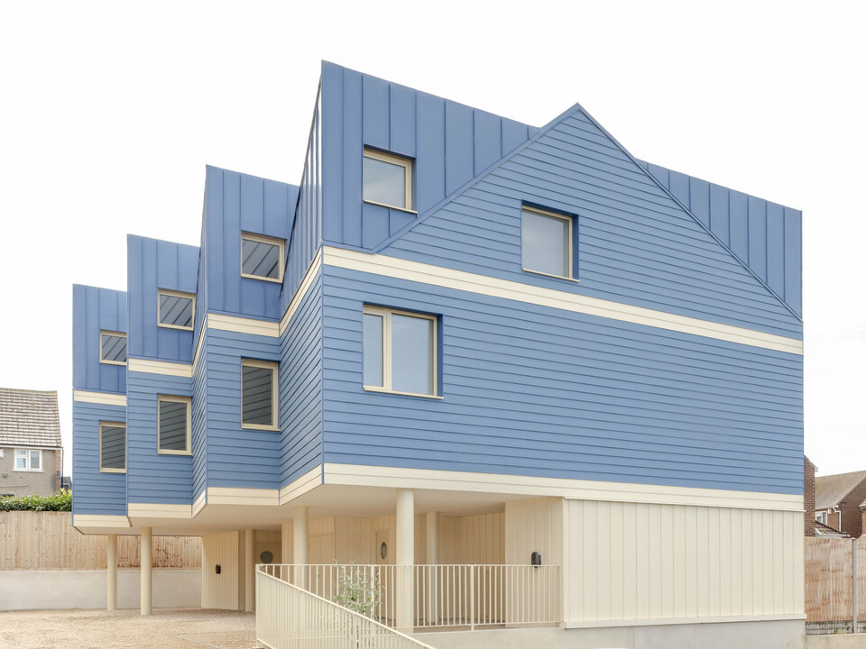 The image shows the striking front of the Goldsmith Mews terraced housing complex with its eye-catching FALZONAL standing seam cladding in dusty blue. The precise, vertical folded panels on the roof and upper floor blend harmoniously with the horizontal aluminium profiles, creating a lively, rhythmic façade. The light-coloured lower areas and the elevated construction lend the building a sense of lightness. The durable, colour-stable PREFA aluminium offers reliable weather protection, high dimensional stability and enables a modern, clear architecture with strong recognition value.