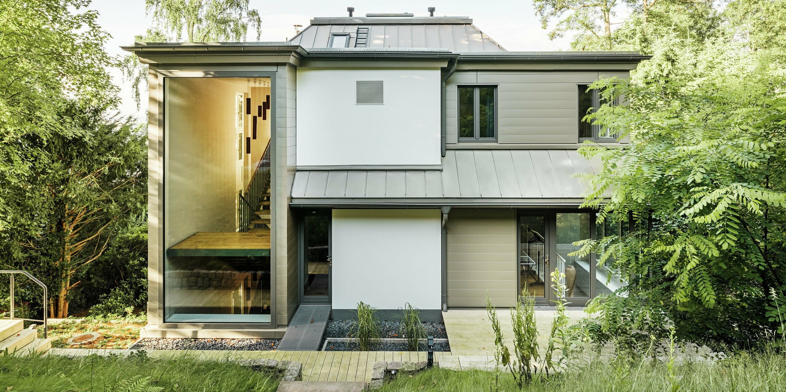 Das Bild zeigt die Rückseite der Villa Blankenese in Hamburg mit einer modernen, klar strukturierten Architektur. Besonders auffällig sind die bronzefarbenen PREFA Sidings, die an der Fassade für eine elegante und zeitlose Optik sorgen. Das Dach ist mit PREFALZ in Bronze ausgeführt und fügt sich harmonisch in das Gesamtbild ein. Große Glasflächen und die Kombination aus Aluminium und Putz unterstreichen den hochwertigen Charakter des Gebäudes. Die langlebigen PREFA Produkte bieten nicht nur ein edles Design, sondern auch maximale Witterungsbeständigkeit und Wartungsfreiheit.