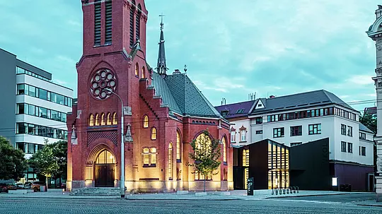 Das Bild zeigt die harmonische Fusion von historischer und moderner Architektur in Olmütz. Die beeindruckende Rote Kirche, mit ihrem markanten spitzen Turm und den gotischen Fenstern, bildet den historischen Mittelpunkt des Ensembles. Direkt daneben erhebt sich der moderne Neubau, dessen Fassade aus matten PREFABOND Aluminium-Verbundplatten und Glas einen klaren, eleganten Kontrast zur traditionellen Architektur bildet. Die großflächigen Glasfronten gewähren Einblicke in den Neubau, der auf innovative Weise mit der Kirche verbunden ist. Der moderne Bau trägt mit seiner präzisen Form und den klaren Linien zur zeitgenössischen Atmosphäre bei, ohne den historischen Charakter der Kirche zu beeinträchtigen. Diese gelungene Symbiose zeigt, wie hochwertig verarbeitete Materialien wie PREFABOND die perfekte Grundlage für die Verschmelzung von Tradition und Moderne bieten.