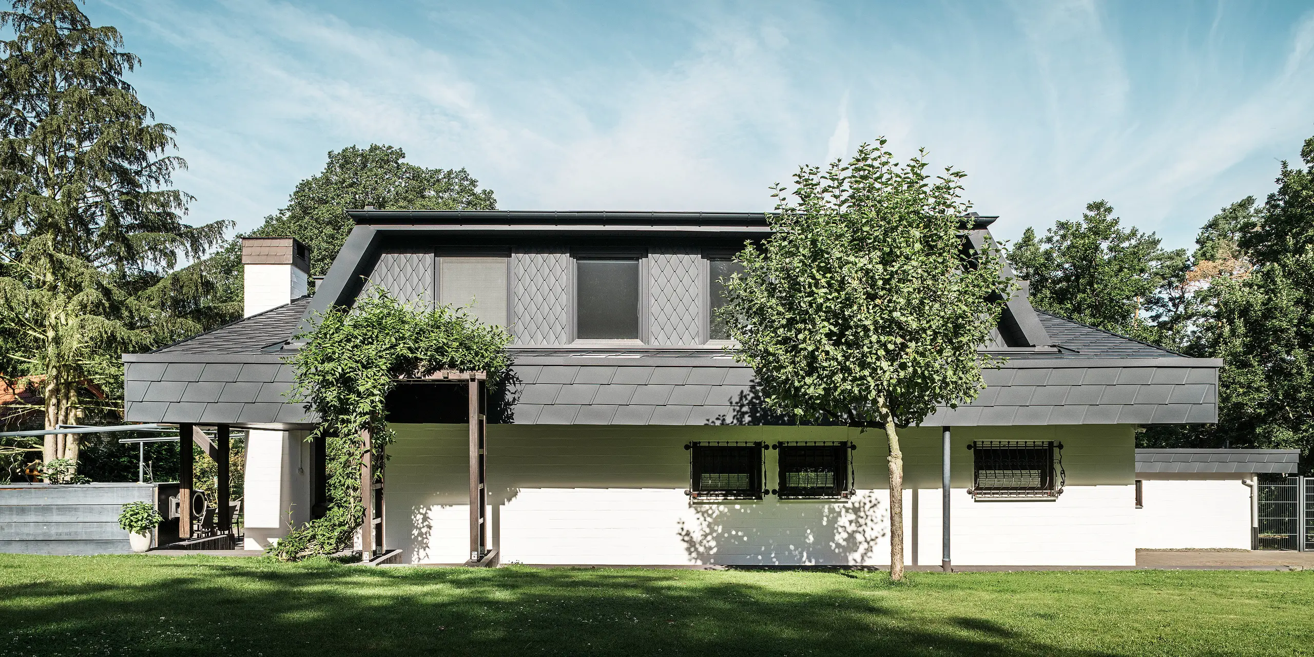 Front view of the garden side of a detached house in Mechernich, featuring a large trapezoidal dormer roof clad with small PREFA P.10 anthracite rhomboid roof tiles. The roof is clad with PREFA DS.19 shingles in the same colour. The façade of the ground floor is painted white, and a green pergola completes the overall look in the garden area. Trees and shrubs frame the view and ensure a harmonious integration into the natural surroundings.