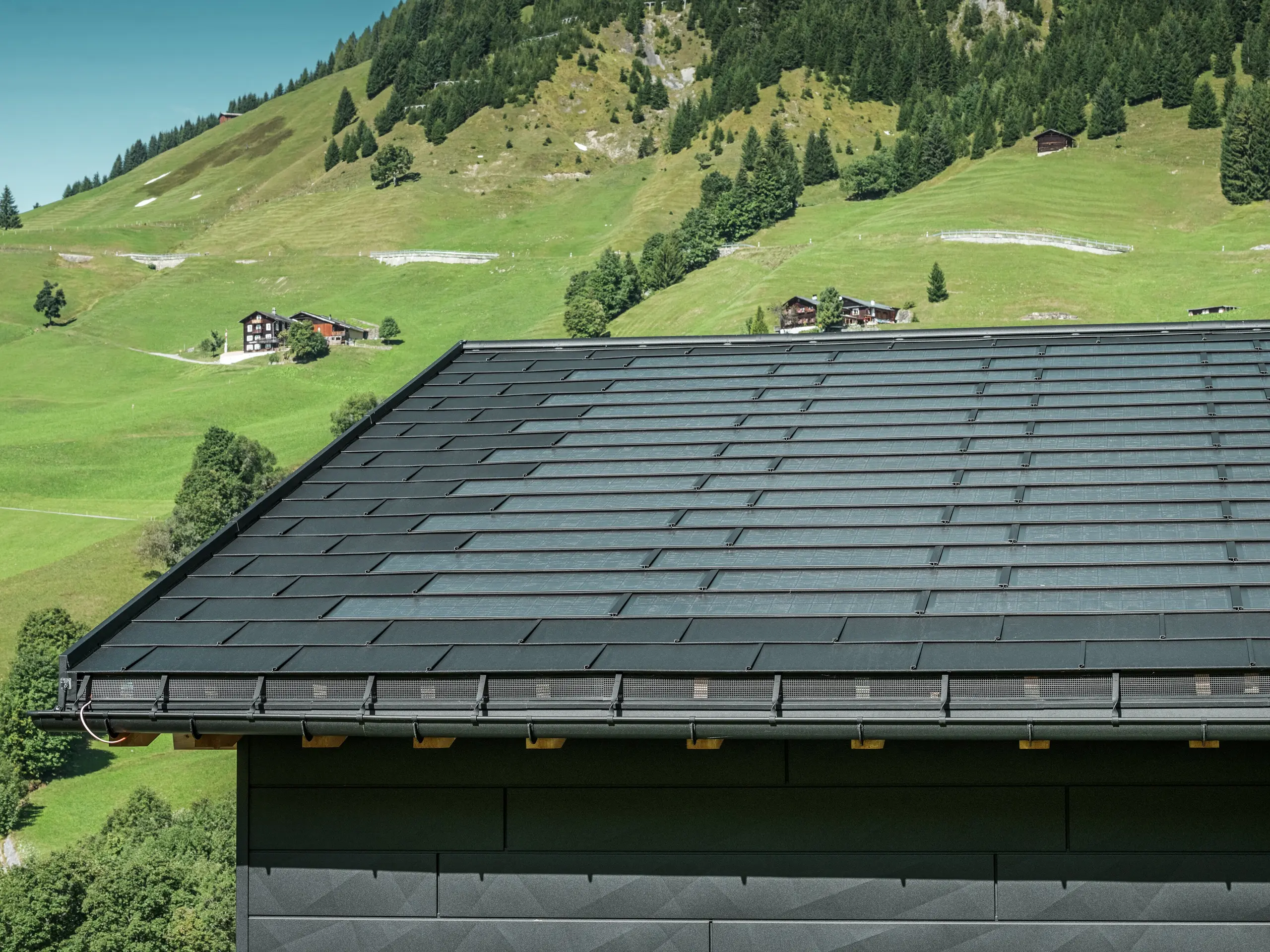 Close-up of the eaves of a modern detached house in the alpine surroundings of Ascharina in Switzerland. The roof was covered with the innovative PREFA solar roof tile and the traditional R.16 roof tile, both in P.10 black. The PREFA half-round gutter and PREFA downpipe ensure reliable roof drainage. The distinctive PREFA Siding.X façade panels can be seen at the bottom of the picture. The clear lines of the aluminium products emphasise the modern design in the midst of a picturesque mountain landscape.