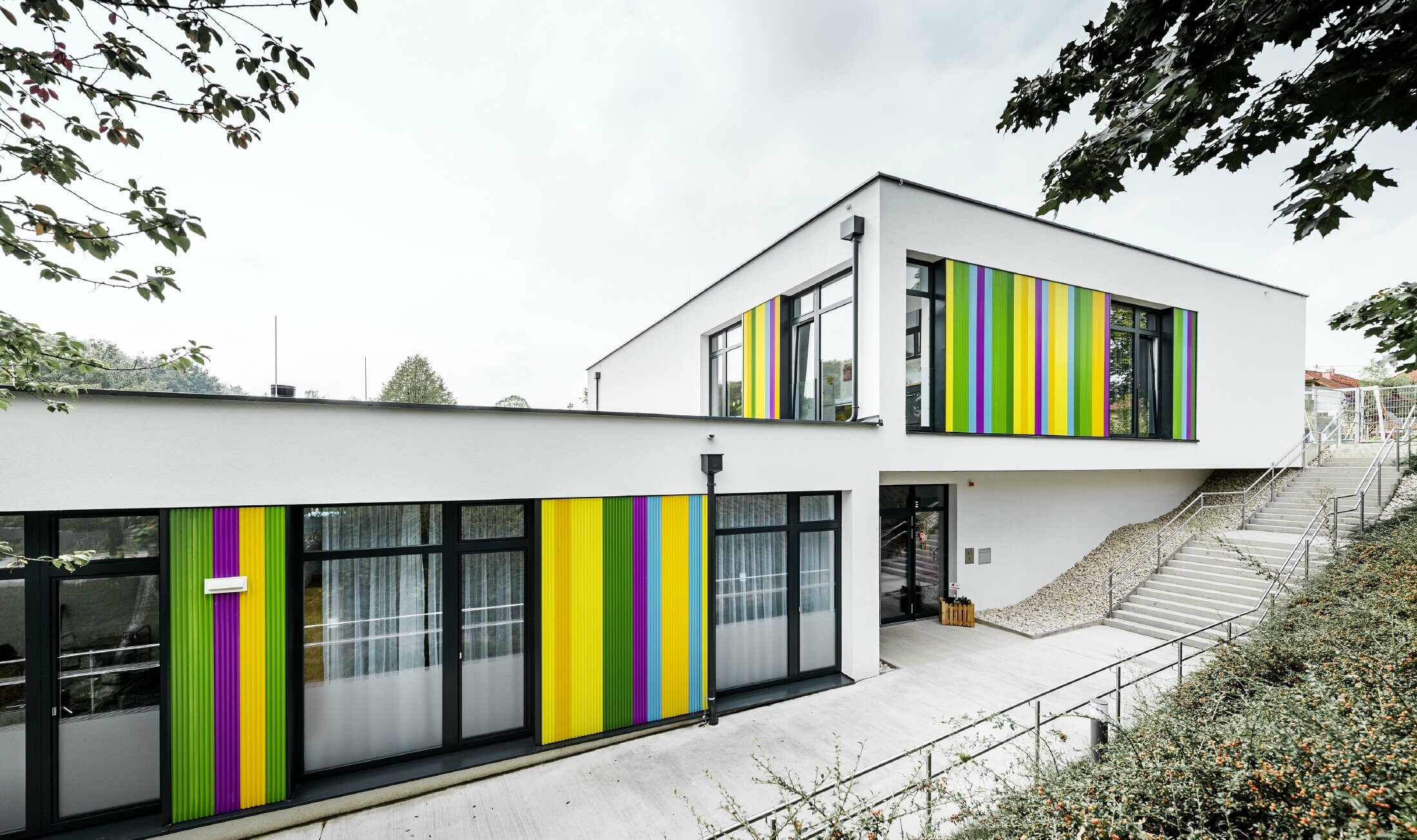 Colourful PREFA façade elements for the kindergarten in Hargelsberg. The building has a flat roof and large, floor-to-ceiling windows.
