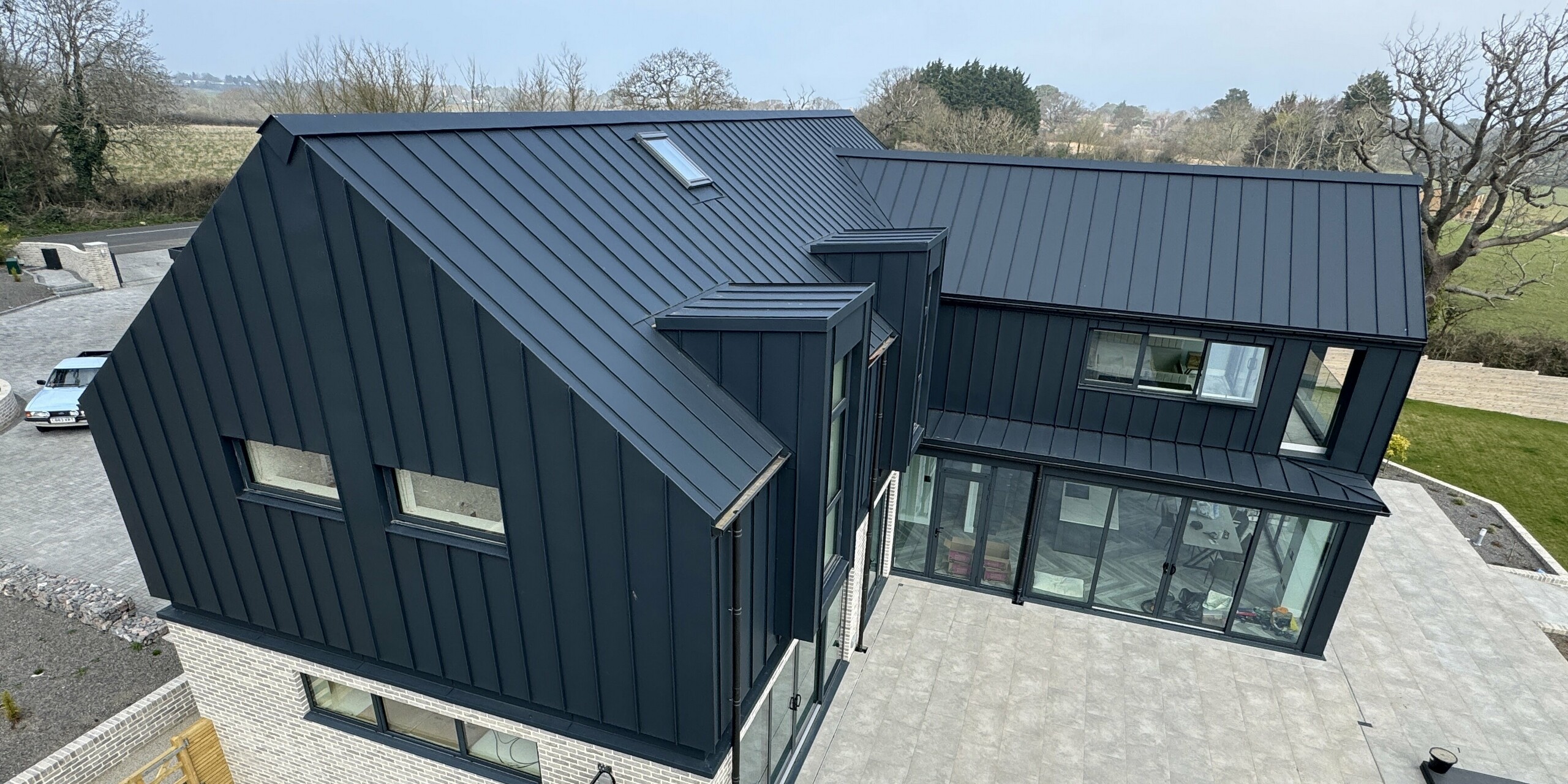 Detailansicht eines modernen Einfamilienhauses mit PREFALZ Dach und Fassade in P.10 Dunkelgrau. Die markante Dachform mit präzisen Stehfalzlinien und die großen Glasflächen prägen das Design. Im Erdgeschoss kontrastiert helle Steinverkleidung mit der dunklen Stehfalzfassade aus Aluminium. Die großzügige Terrasse aus grauen Platten umrahmt das Gebäude und öffnet den Blick auf den Garten. PREFALZ überzeugt durch Langlebigkeit, Korrosionsbeständigkeit und Wartungsfreiheit – ideal für die exponierte Lage auf der Isle of Wight.