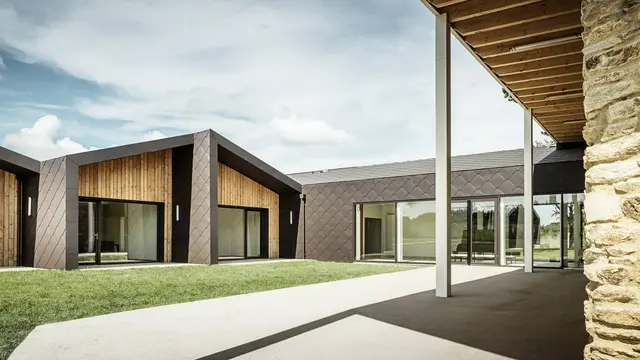 Das Bild zeigt ein modernes Gebäude mit einer Fassade aus PREFA 44 × 44-Dach- und Wandelementen in Walnussbraun. Die Fassade besticht durch ihre elegante Rautenstruktur, die sich harmonisch in die warmen Holzelemente einfügt. Große Glasflächen lassen viel Tageslicht ins Innere und schaffen eine offene und luftige Atmosphäre. Ein überdachter Durchgang auf der rechten Bildseite führt zu einer rustikalen Steinmauer, die einen interessanten Kontrast zum modernen Design bildet.