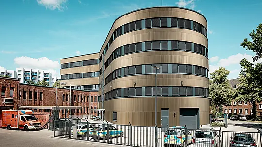 Markante Außenansicht des Polizeikommissariats 43 in Hamburg: Der geschwungene Neubau mit PREFA Aluminiumfassade aus eloxierten Strangpressprofilen „Zacke“ in Mittelbronze wirkt als kraftvoller Eyecatcher im Stadtbild. Die feine, vertikale Struktur erzeugt lebendige Licht- und Schatteneffekte und betont die rund geformte Architektur. Im Kontrast dazu steht der historische Backsteinbestand. PREFA Aluminium überzeugt durch hohe Witterungsbeständigkeit, Korrosionsschutz, Langlebigkeit und minimalen Wartungsaufwand – ideal für nachhaltige, funktionale und ästhetische Fassadenlösungen.