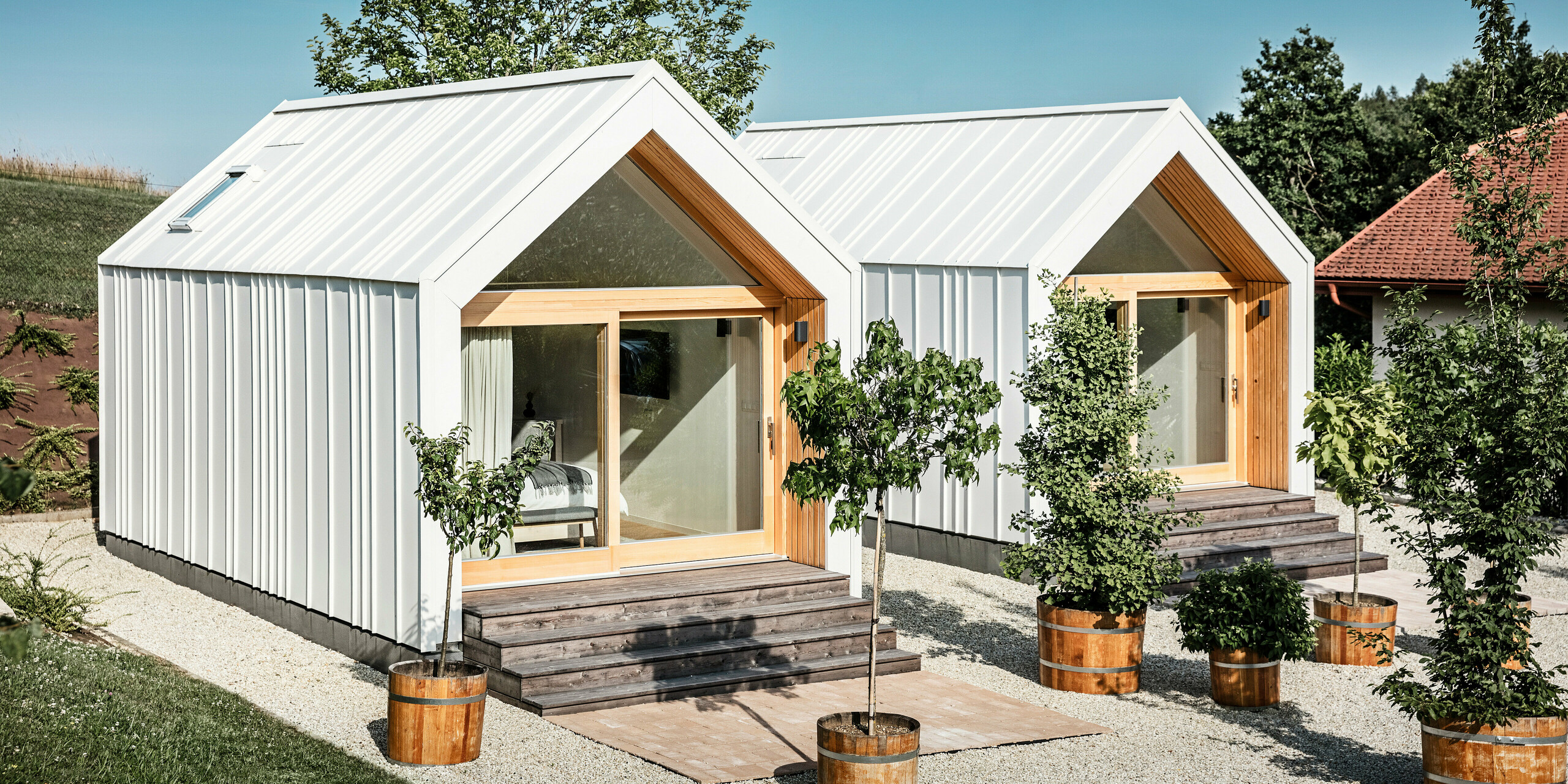 Front view of the two Tiny Houses of the 'Pri Momi' holiday complex in Kančevci, Slovenia. The exclusive guest accommodation was clad with robust PREFALZ roof and façade cladding in the colour P.10 Pure White. The modern micro houses combine an aesthetic and durable aluminium shell with large glass fronts and natural wood, creating an inviting and warm atmosphere. The surroundings are characterised by manicured lawns and small trees in planters, which complement the minimalist design of the houses.