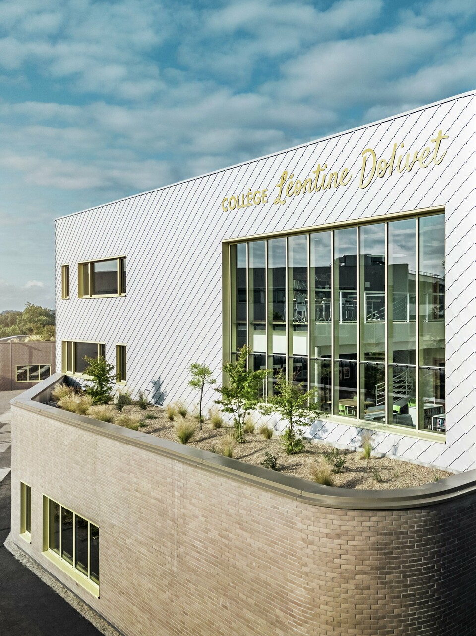Das Foto zeigt eine markante Gebäudeecke des Collège Léontine Dolivet in Cesson-Sévigné. Die Fassade besteht aus silbermetallic PREFA Wandrauten, die in einem diagonalen Muster angeordnet sind und das Licht reflektieren. Auffällig ist die geschwungene Form der oberen Etage, die dem Bau eine weiche, moderne Anmutung verleiht. Große Glasfronten öffnen den Blick ins Innere, während gelbe Fensterrahmen lebendige Akzente setzen. Im Vordergrund befindet sich ein bepflanzter Bereich mit Kies und Sträuchern, der die Architektur harmonisch ergänzt. Der Himmel ist leicht bewölkt, was die metallische Oberfläche betont.