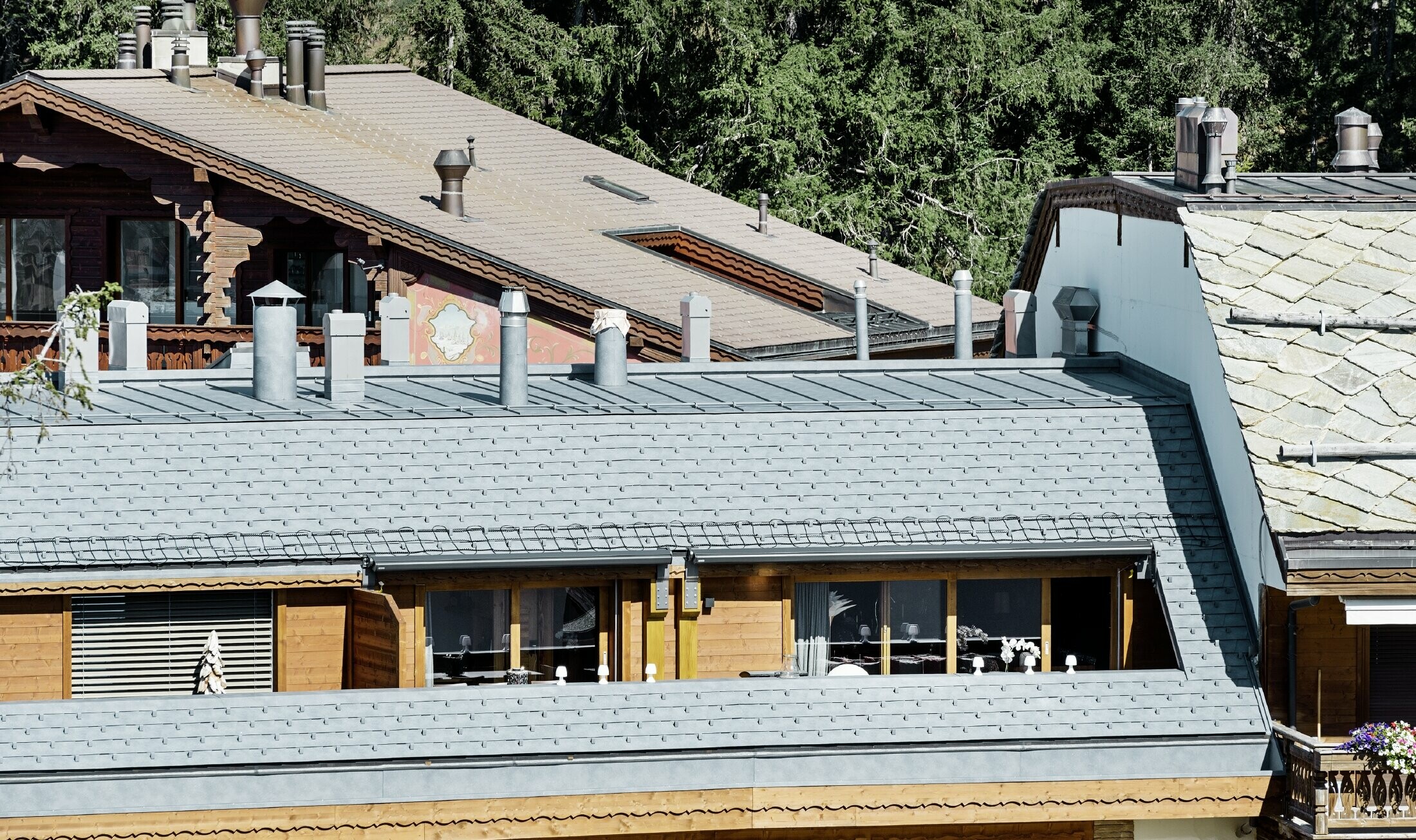 Apartment building in Crans-Montana (Switzerland) with mountains in the background, a façade with lovely wooden elements and a PREFA aluminium shingle roof in stone grey