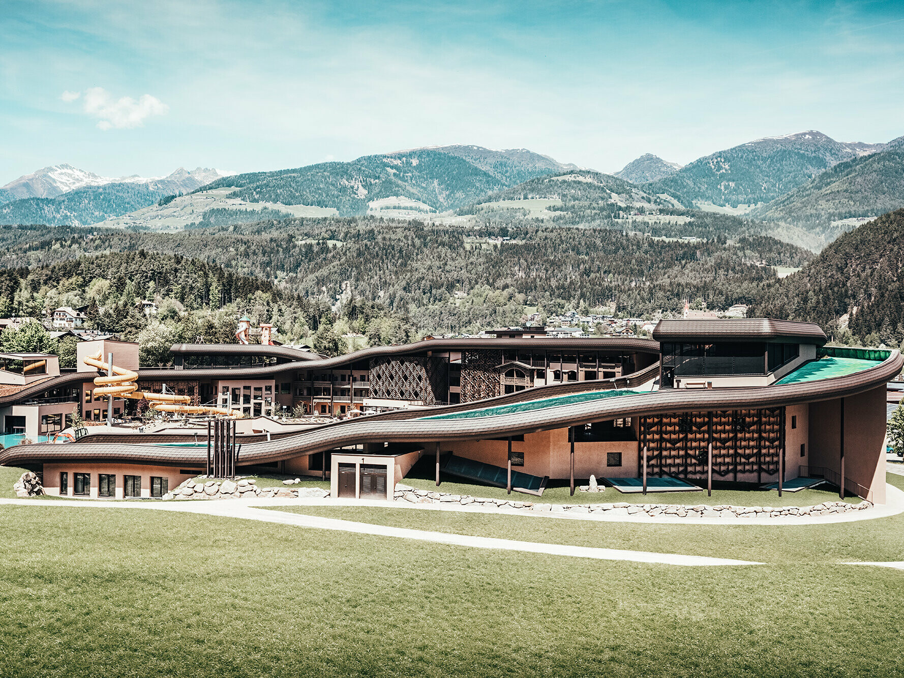 View of the entire hotel complex from a distance: The roof wave runs across the entire building with a beautiful  hilly, wooded landscape in the background