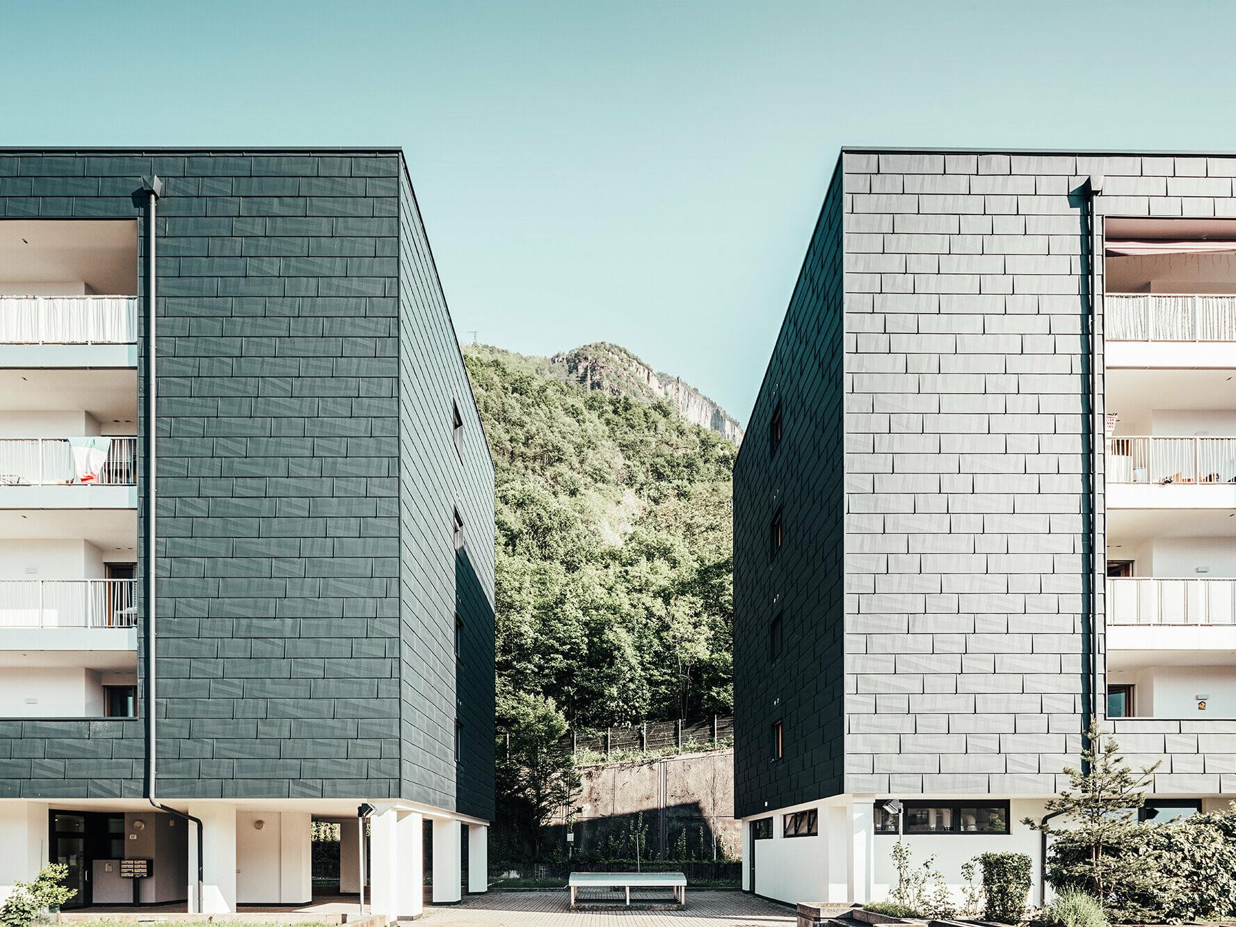 Photo of parts of two apartment blocks with the anthracite-colored PREFA façade. In between, there are wooded mountains in the background.