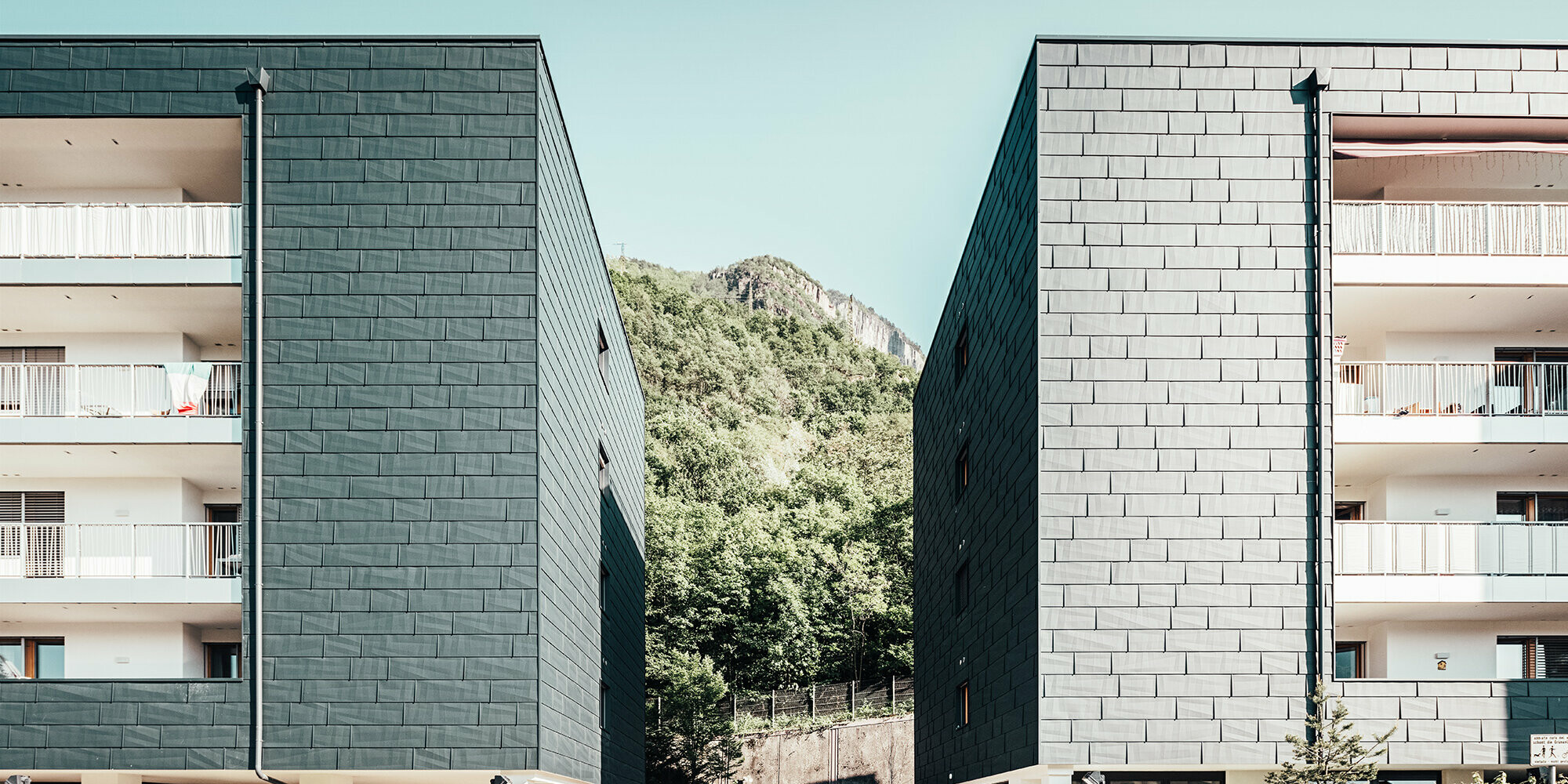 Photo of parts of two apartment blocks with the anthracite-colored PREFA façade. In between, there are wooded mountains in the background.