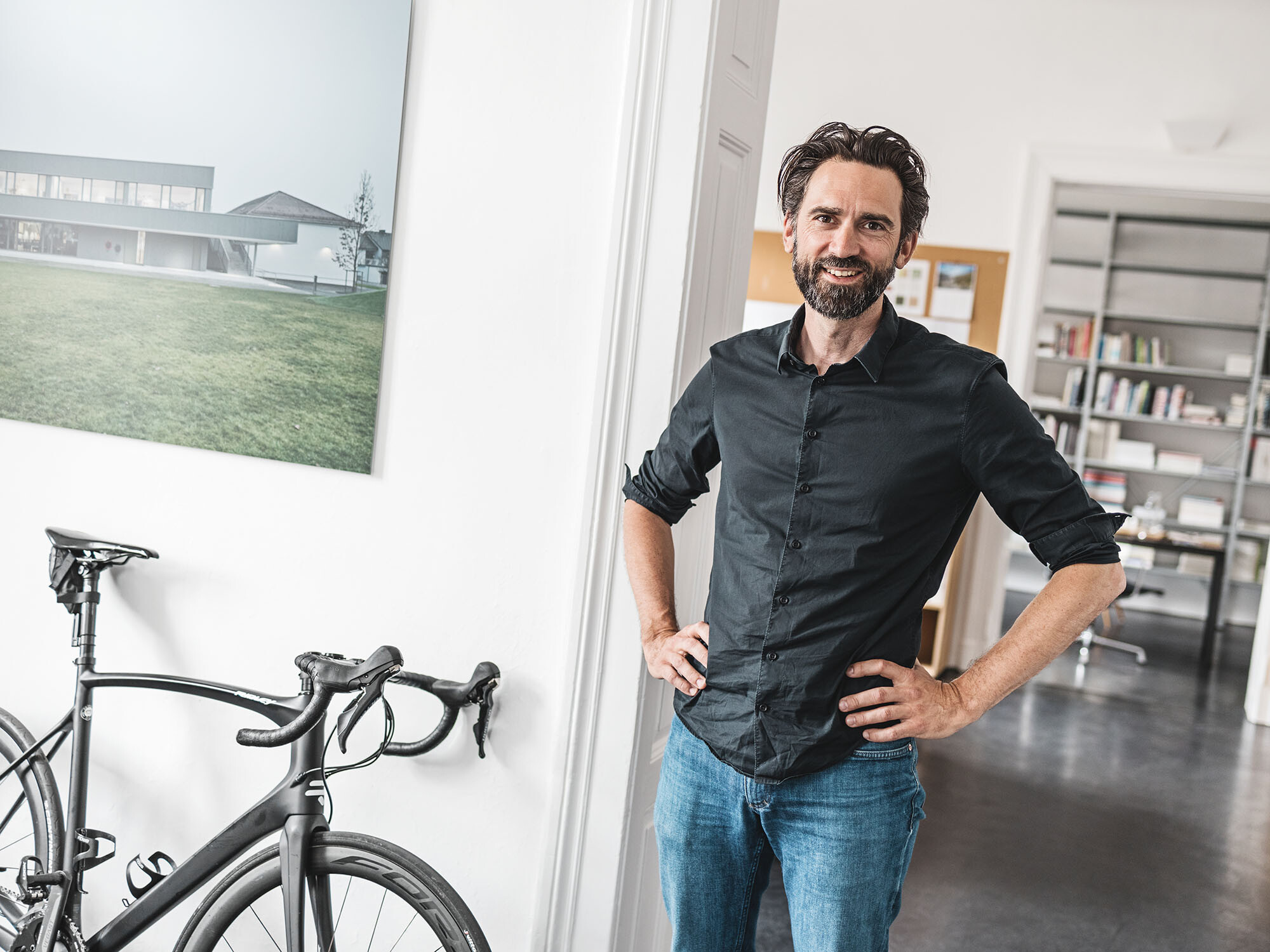 A portrait of the lead architect Thomas Heil, an office space extending behind him and a bicycle leaning against the wall next to him.