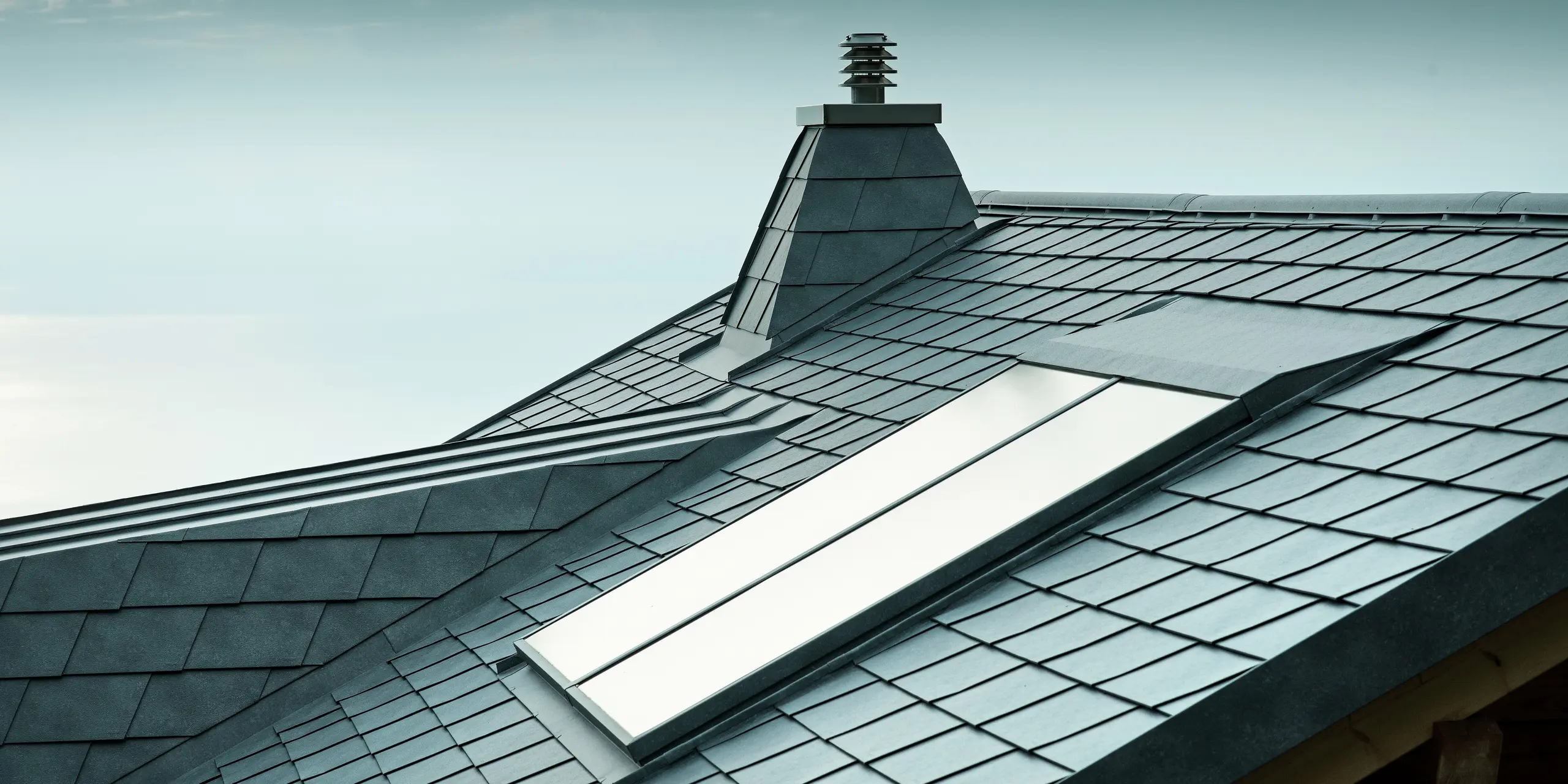 Diese vielseitige Dachansicht zeigt steingraue PREFA-Dachschindeln sowie einen Kamin in Trapezform aus Aluminium. Das langlebige Aluminiumdach ist nicht nur stilvoll, sondern auch äußerst widerstandsfähig. Die exakte Ausführung der Dachgaube und des Dachfensters zeugt von der Detailgenauigkeit der PREFA-Verlegepartner.