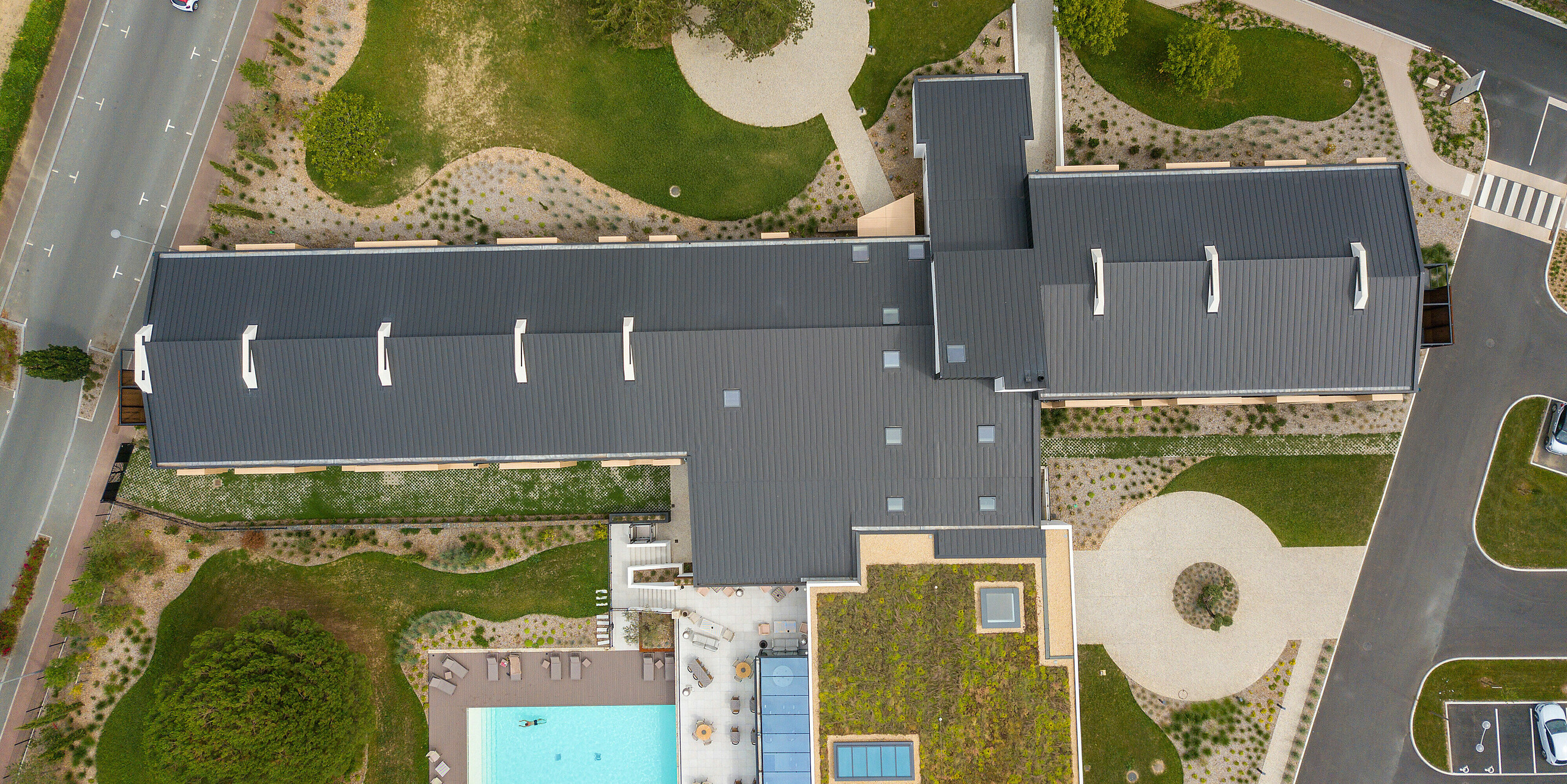 Holistische Drohnenaufnahme auf des Hôtel Parc du Landreau in Les Herbiers, Frankreich, mit PREFALZ Dach in Schwarzgrau. Das langlebige Aluminium-Stehfalzdach bietet optimalen Schutz vor Witterungen und fügt sich harmonisch in das architektonische Konzept des Hotels ein. Der Außenbereich mit Swimmingpool und begrüntem Dach zeigt die moderne und nachhaltige Bauweise.