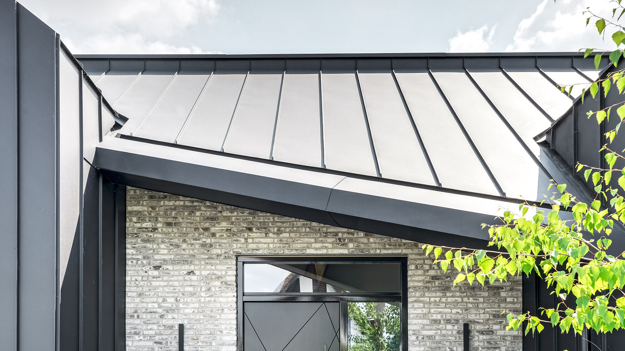 Detailed view of a modern building with a steeply pitched, black prefabricated roof. The metal seam roof covers an elegant façade with large glass windows and a brick wall. The black metal frame around the window contrasts with the texture of the light-coloured bricks and the roof blends harmoniously into the overall picture. Green leaves in the foreground create natural accents.