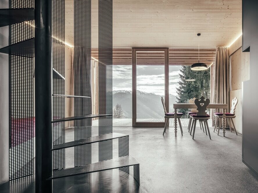 Interior shot. To be seen: the spiral staircase, the exposed concrete floor, rustic carpentry furniture. View over the spacious balcony towards the Dolomites.