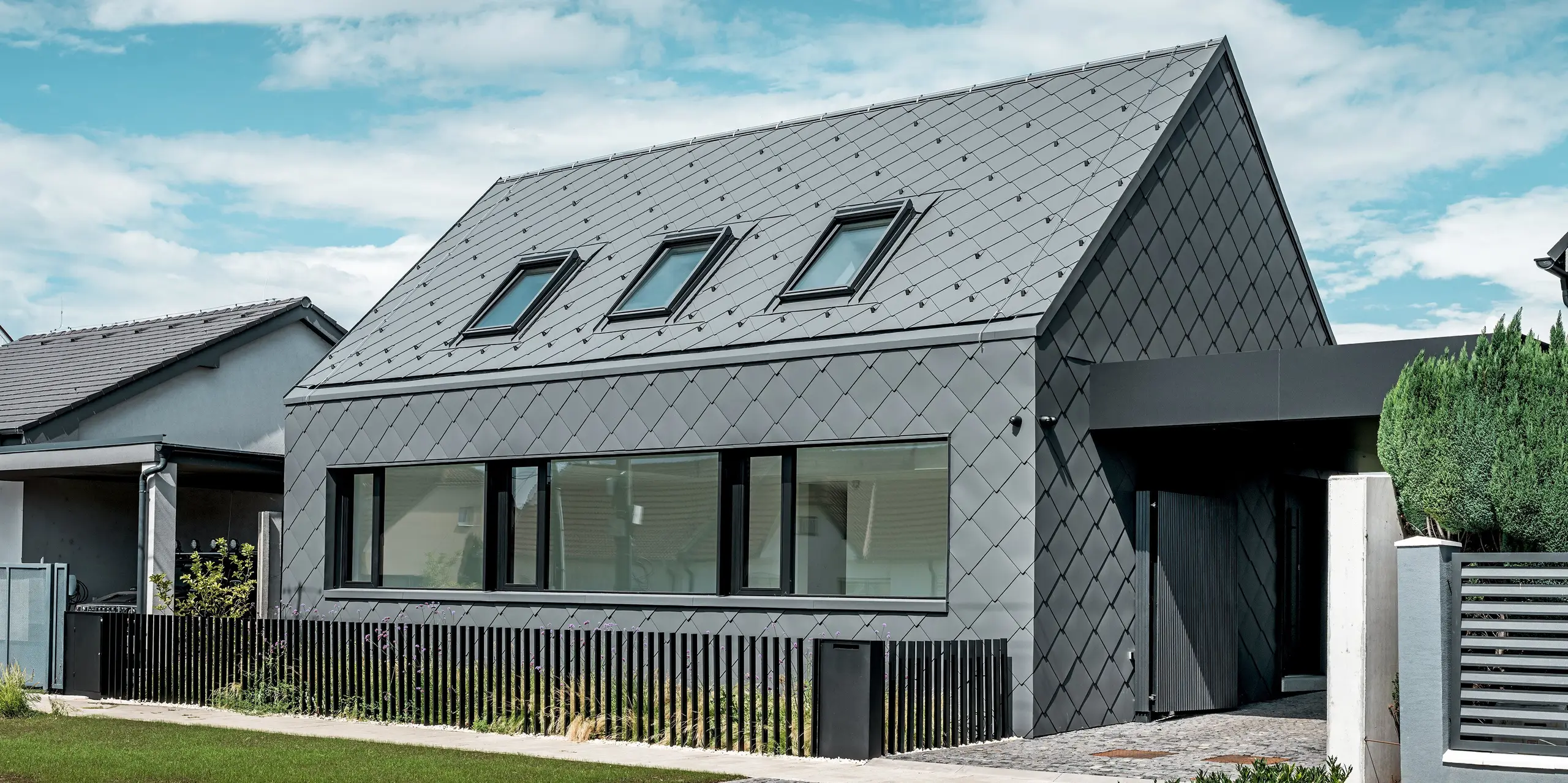 Modernes Einfamilienhaus in Trnava‑Modranka mit Aluminiumhülle: Die gesamte Gebäudehülle dieses schlichten Gebäudes wurde mit PREFA Dach- und Wandrauten 44 × 44 in P.10 Dunkelgrau gestaltet. Die markante Rautenstruktur zieht sich nahtlos über Dach und Fassade und verleiht dem Haus eine starke, monolithische Wirkung. Große Fensteröffnungen setzen klare architektonische Akzente, während das witterungsbeständige PREFA Aluminium langlebigen Schutz und eine edle, matte Optik bietet. Der helle Himmel verstärkt den Kontrast zur dunklen Gebäudehülle und betont das moderne Design.