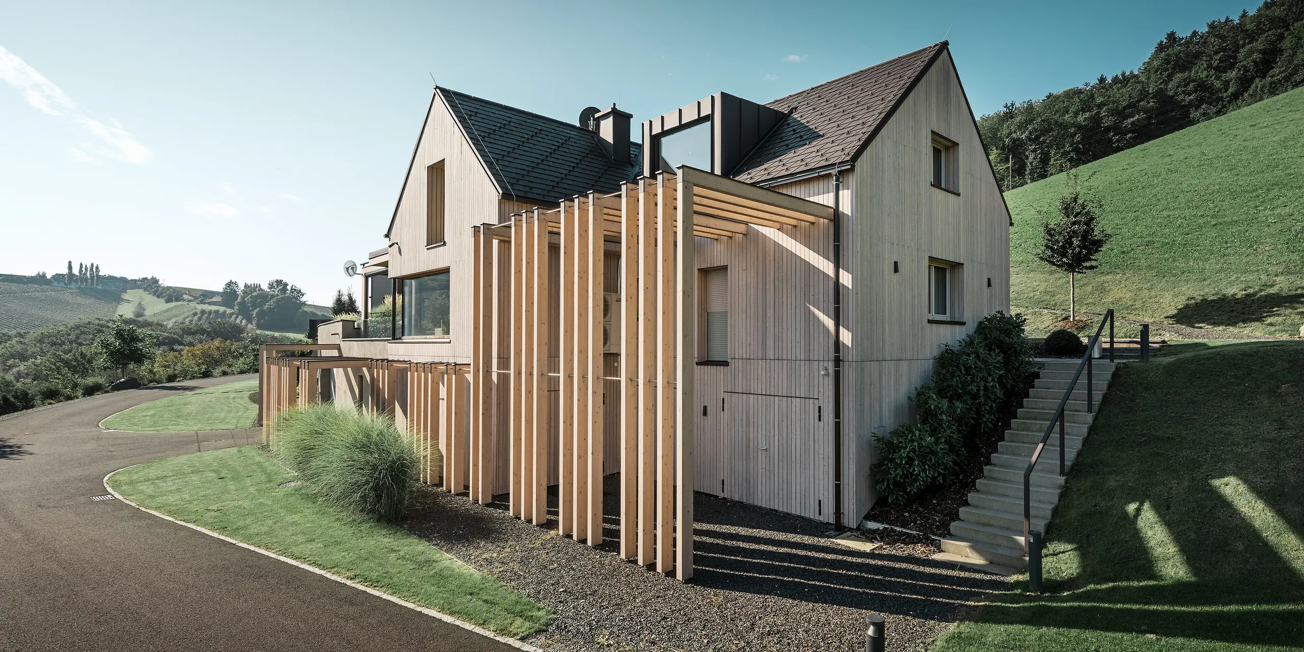 Large detached house with gabled roof and dormer window on a hillside. The roof is covered with brown PREFA aluminium shingles and the façade is clad with vertical wooden boards.