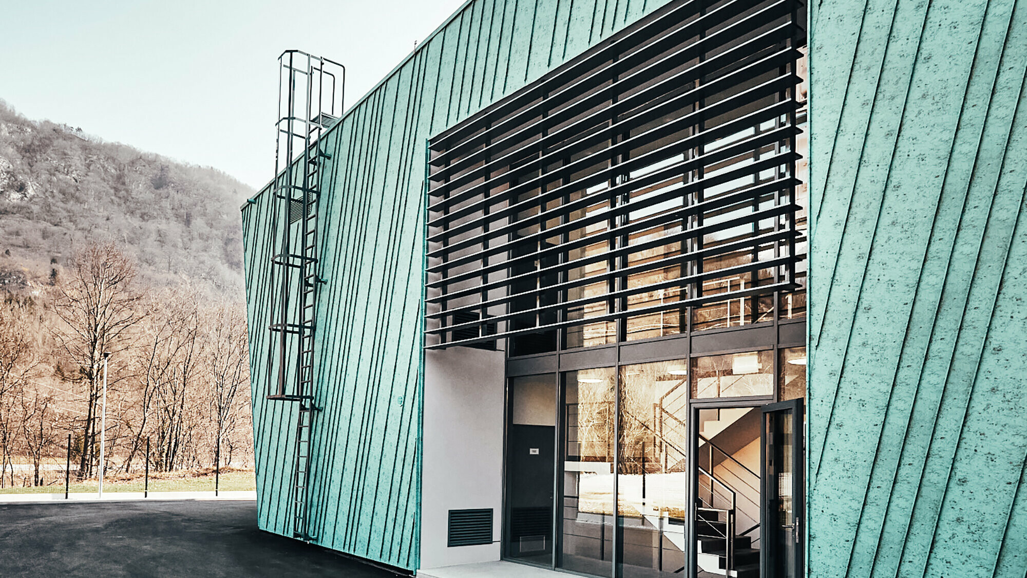 Detail with slightly slanted façade and a small, elongated window that reflects the mountains and electrical lines. Lateral view of the façade and glass front with an entrance. 