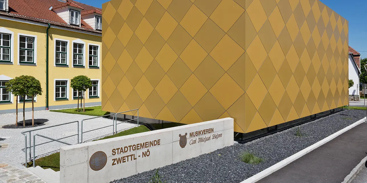 Eingangsbereich zum Musikverein Zwettl. Vor dem Gebäude ist die Funktion des Objekts in Stein gemeißelt. Das quaderförmige Gebäude besticht durch die markante Optik mit PREFA Aluminium Verbundplatten in zwei unterschiedlichen Goldtönen. Die Aluminiumplatten sind sorgfältig in Form von Karos auf der Fassade montiert worden. 