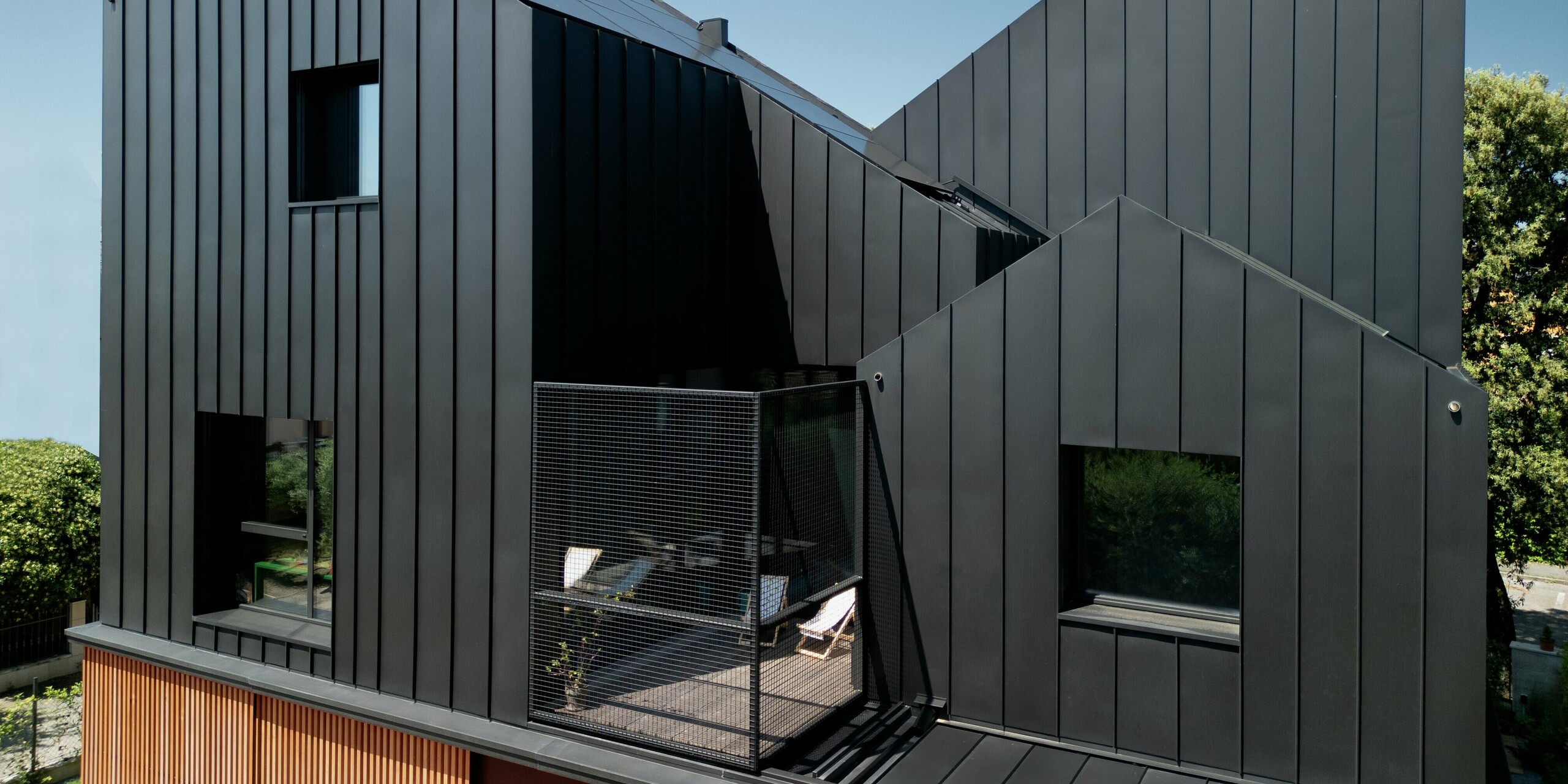 Gesamtansicht des Obergeschosses des modernen Doppelhauses in Cattolica: Markante Aluminiumfassade und Dach mit PREFALZ in Schwarzgrau, kombiniert mit warmer Holzverkleidung. Die klare Linienführung und die hochwertige PREFA Oberfläche sorgen für Langlebigkeit, Korrosionsschutz und ein elegantes, zeitloses Design unter blauem Himmel.