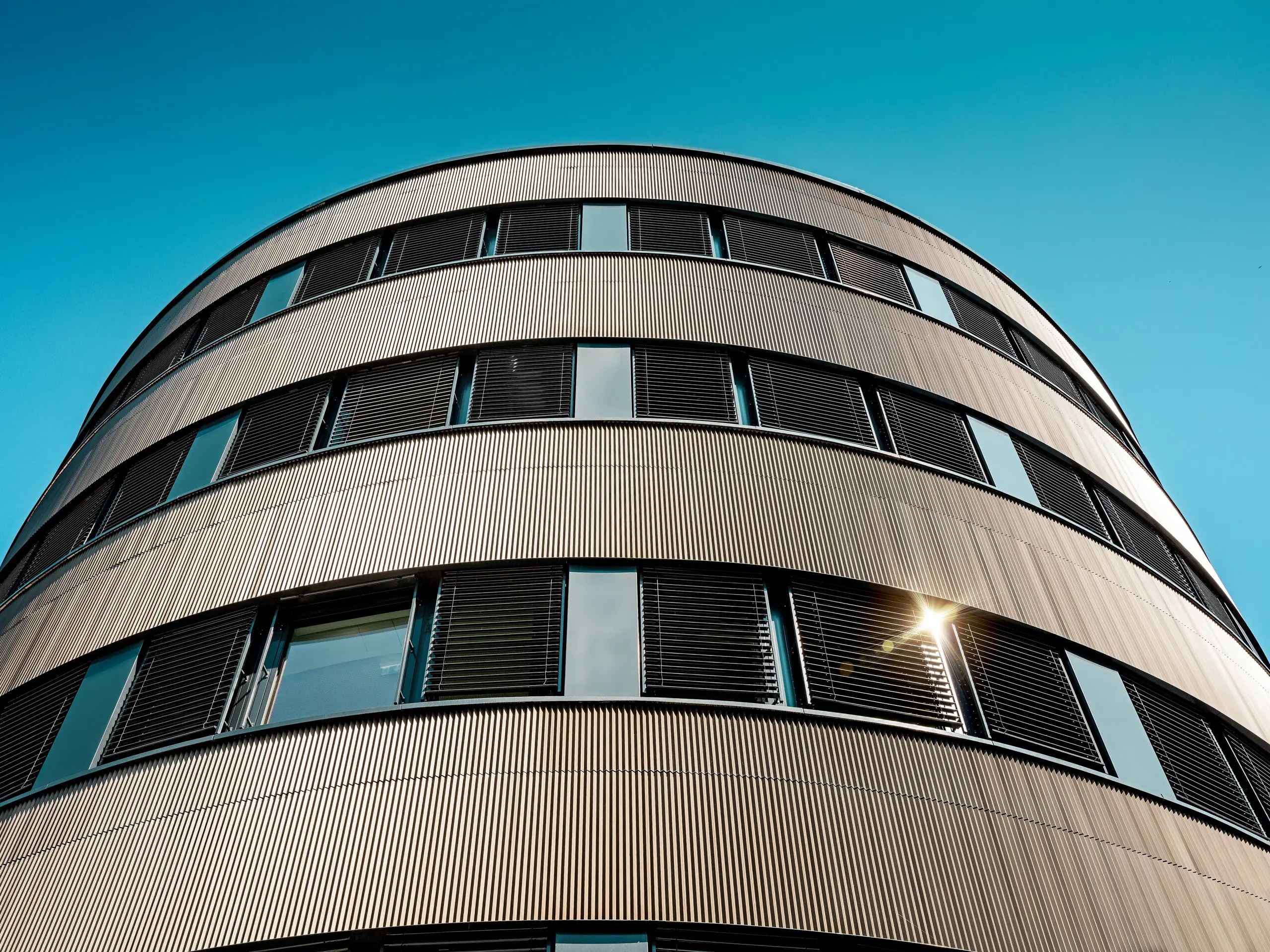Dramatische Untersicht des geschwungenen Neubaus des Polizeikommissariats 43 in Hamburg: Die PREFA Aluminiumfassade aus eloxierten Strangpressprofilen „Zacke“ in Mittelbronze entfaltet als Eyecatcher ihre volle Wirkung. Die fein geriffelte Struktur reflektiert das Sonnenlicht in lebendigen Nuancen und unterstreicht die präzisen Rundungen sowie die horizontalen Fensterbänder. PREFA Aluminium überzeugt dabei durch hohe Witterungsbeständigkeit, Korrosionsschutz, Langlebigkeit und minimalen Wartungsaufwand – ideal für nachhaltige, architektonisch anspruchsvolle Fassadenlösungen.