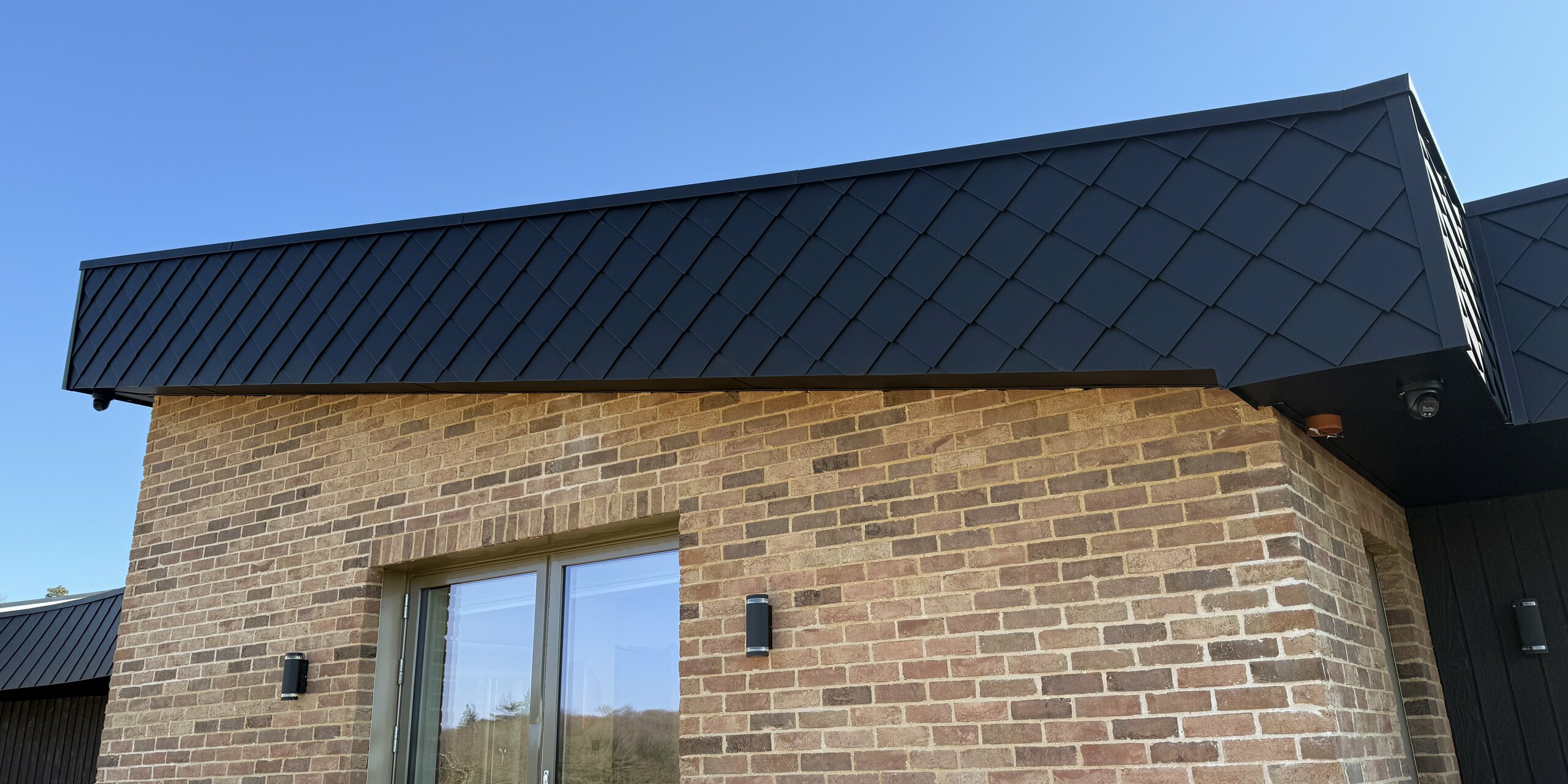 Modernes Einfamilienhaus mit markanter Fassadengestaltung: Die Kombination aus warmen Klinkern und der eleganten PREFA Wandraute 20 × 20 in der Farbe P.10 Anthrazit setzt starke Akzente an der Hausfassade. Die robusten Blechrauten verleihen dem Gebäude ein exklusives Design und überzeugen durch Langlebigkeit, Witterungsbeständigkeit und Pflegeleichtigkeit. Vor strahlend blauem Himmel kommt die geometrische Struktur besonders gut zur Geltung und unterstreicht die hochwertige Architektur.