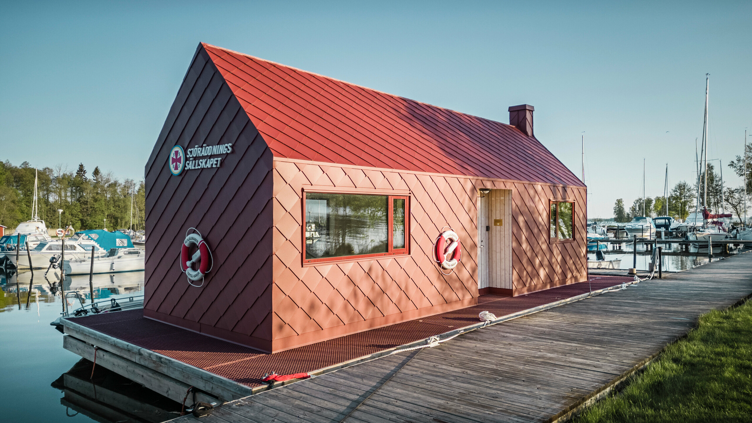 Die Seenotrettungsstation am Hjälmaren See in Schweden, festgemacht an einem Holzsteg. Das markante Gebäude mit einer Fassade aus oxydroten PREFA Dach- und Wandrauten 29×29 steht stolz im Kontrast zu den weißen und blauen Booten im Hintergrund. Mit Rettungsringen ausgestattet und bereit für den Einsatz, vermittelt das Haus sowohl Sicherheit als auch einen starken maritimen Charakter in dieser friedvollen Seenlandschaft.