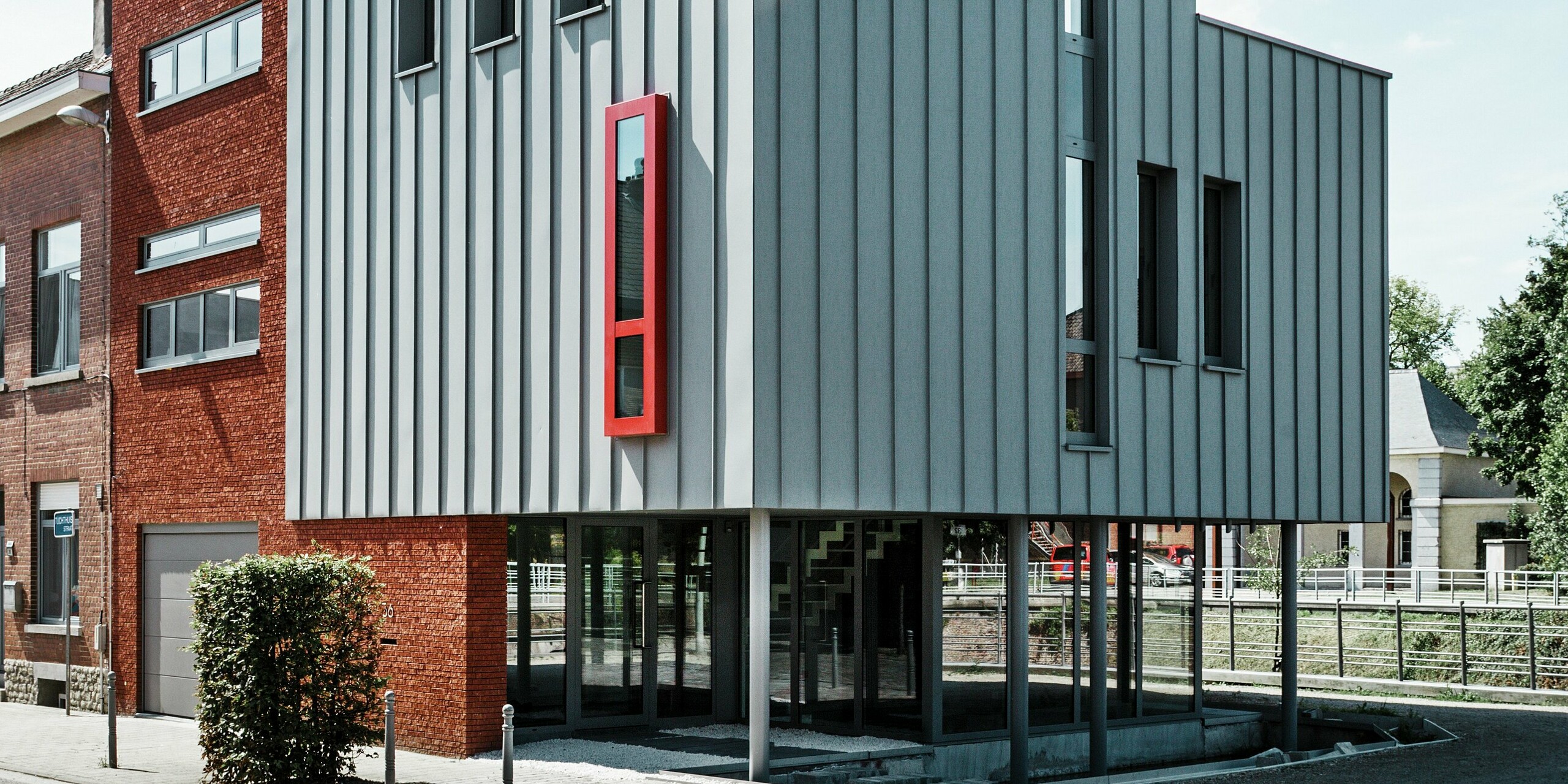 Modernes Wohn- und Bürogebäude in Vilvoorde, Belgien, mit markanter PREFALZ Aluminiumfassade in Patinagrau. Die vertikale Stehfalzstruktur sorgt für eine elegante Linienführung, während das leuchtend rote Fenster zum Eyecatcher wird. Großflächige Verglasungen und ein schwebend wirkendes Erdgeschoss verleihen dem Bau Leichtigkeit und architektonische Raffinesse.