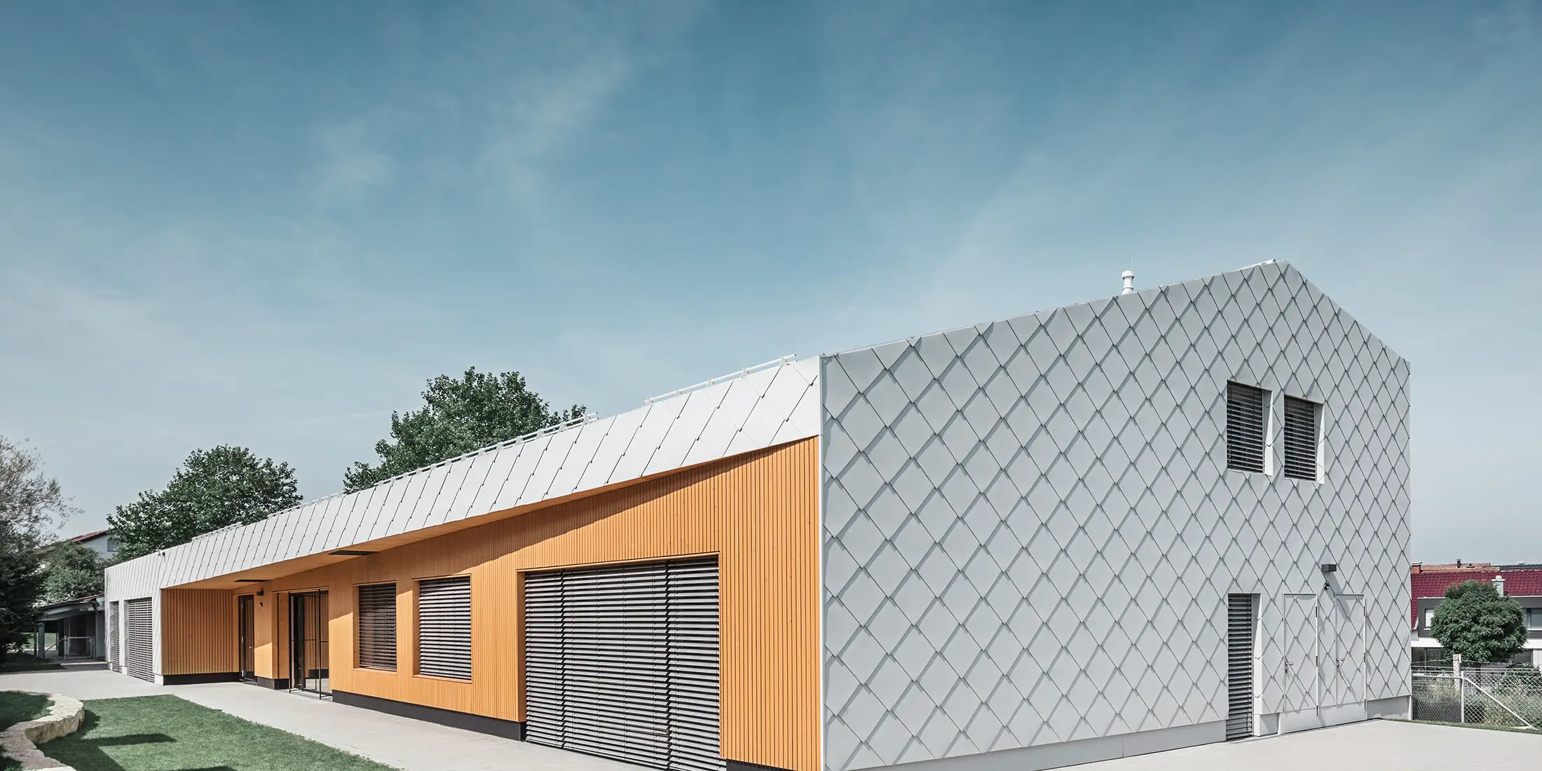 Modernes Kindergartengebäude mit Kombination aus silberner Aluminiumrautenfassade und Holzverkleidung in warmem Orangeton. Die Architektur vereint klare Linien mit funktionalen Elementen wie großen Fenstern und Sonnenschutzlamellen. Das Gebäude steht auf einem gepflasterten Gelände mit angrenzender Rasenfläche unter blauem Himmel.