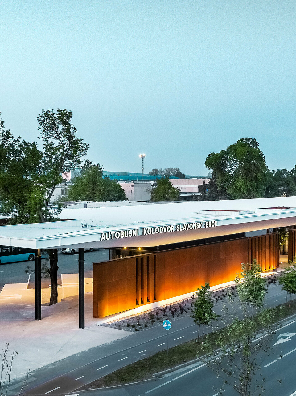 The picture shows the bus stop "Autobusni Kolodvor Slavonski Brod" in Croatia at dusk. The bus stop has a large, white Prefalz roof from PREFA, which is supported by black columns. The façade consists of modern, brown panels, which are illuminated by floor lights, creating a warm, inviting ambience. Buses and waiting areas can be seen under the roof. In the background are various buildings, including a tall office tower and several residential buildings. 