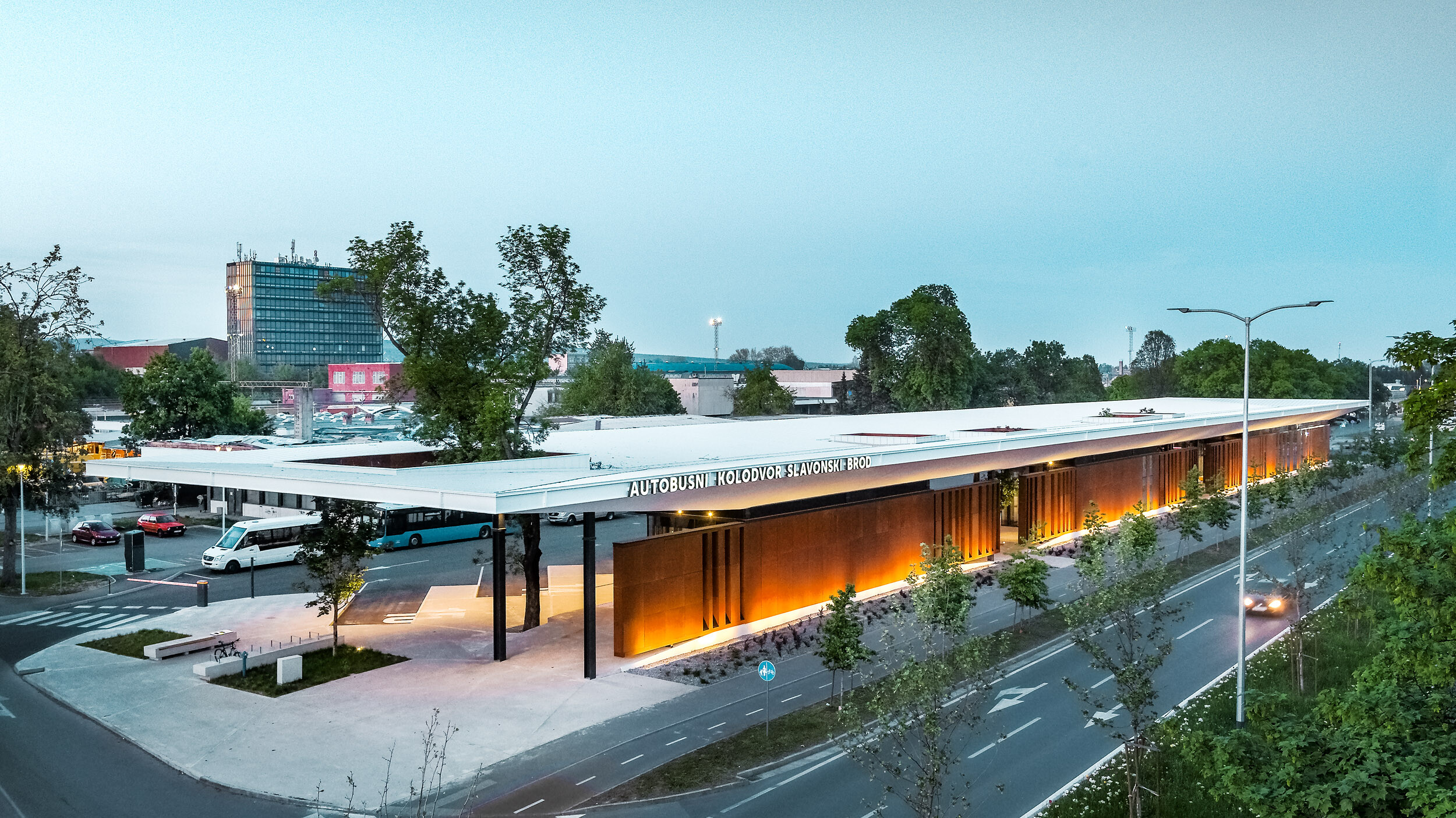 The picture shows the bus stop "Autobusni Kolodvor Slavonski Brod" in Croatia at dusk. The bus stop has a large, white Prefalz roof from PREFA, which is supported by black columns. The façade consists of modern, brown panels, which are illuminated by floor lights, creating a warm, inviting ambience. Buses and waiting areas can be seen under the roof. In the background are various buildings, including a tall office tower and several residential buildings. 