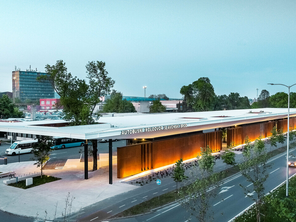 The picture shows the bus stop "Autobusni Kolodvor Slavonski Brod" in Croatia at dusk. The bus stop has a large, white Prefalz roof from PREFA, which is supported by black columns. The façade consists of modern, brown panels, which are illuminated by floor lights, creating a warm, inviting ambience. Buses and waiting areas can be seen under the roof. In the background are various buildings, including a tall office tower and several residential buildings. 