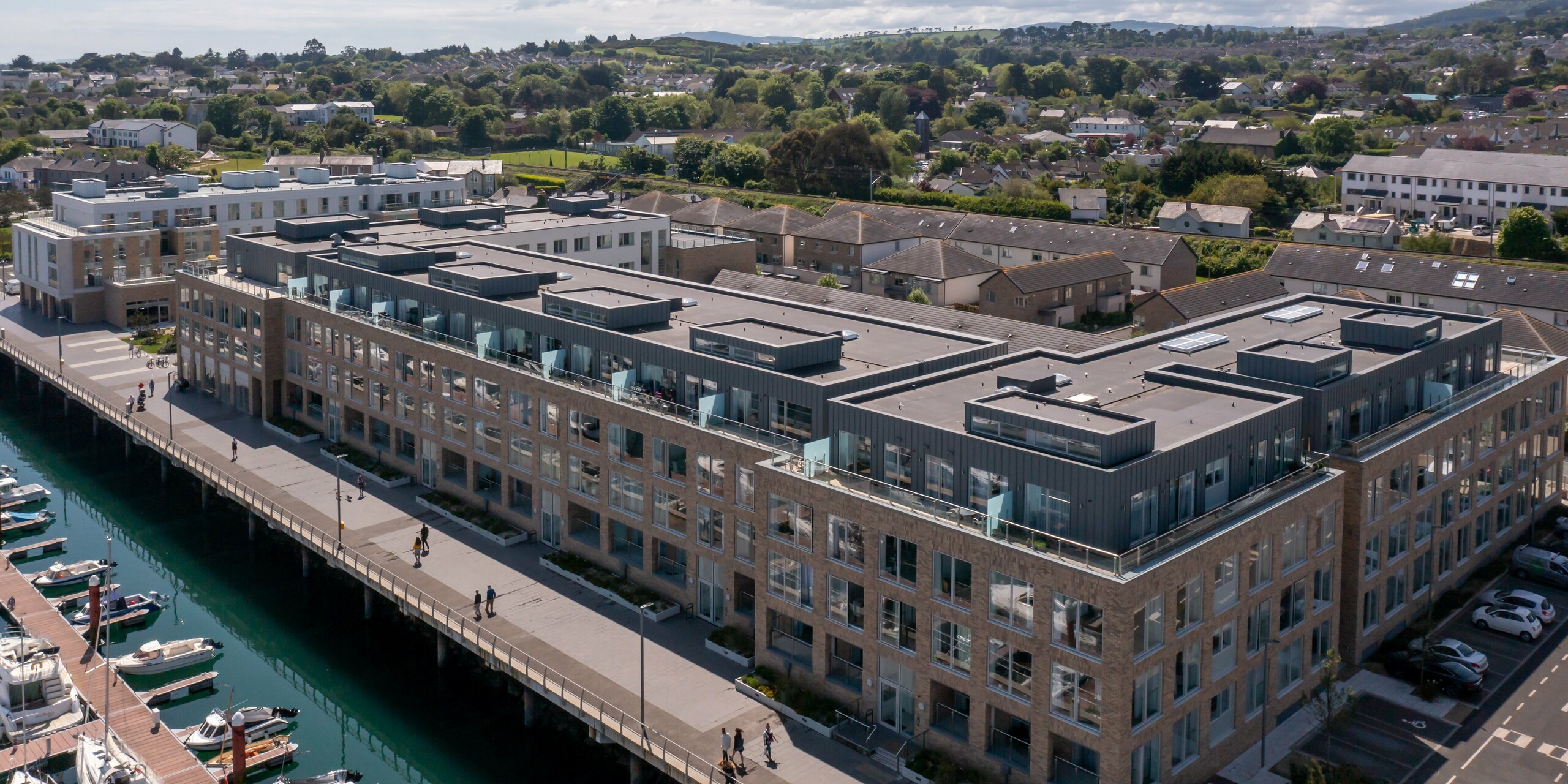 Wide-angle bird's eye view of Marina Village Greystones in Wicklow, Ireland - a modern waterfront residential development directly on the marina. The building complex is clad with robust PREFALZ in P.10 Prefa White and P.10 Light Grey. The elegant architecture includes large glass fronts, spacious balconies and a high quality sheet metal façade design. The use of PREFALZ aluminium provides durable protection and an attractive appearance. Surrounded by a picturesque landscape, the residential project represents exclusive living by the sea.
