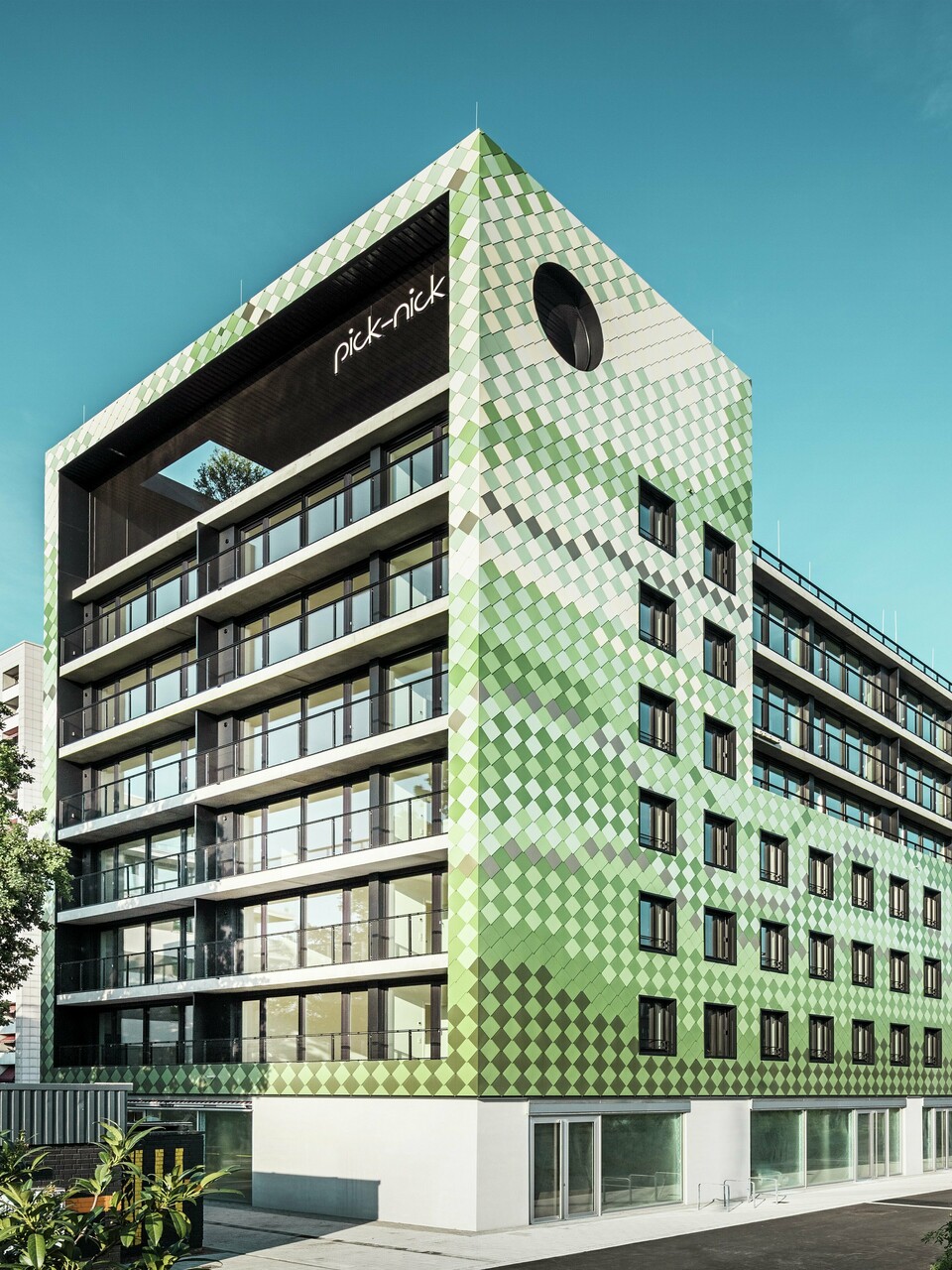 The ‘Pick-Nick’ residential and commercial building in Dresden boasts a modern PREFA aluminium façade made of rhomboid panels in various shades of green and beige. The striking building envelope combines clear lines with a dynamic play of colours and is complemented by generous glass surfaces. The ‘Pick-Nick’ lettering on the upper edge of the building and the green roof terrace add architectural highlights.