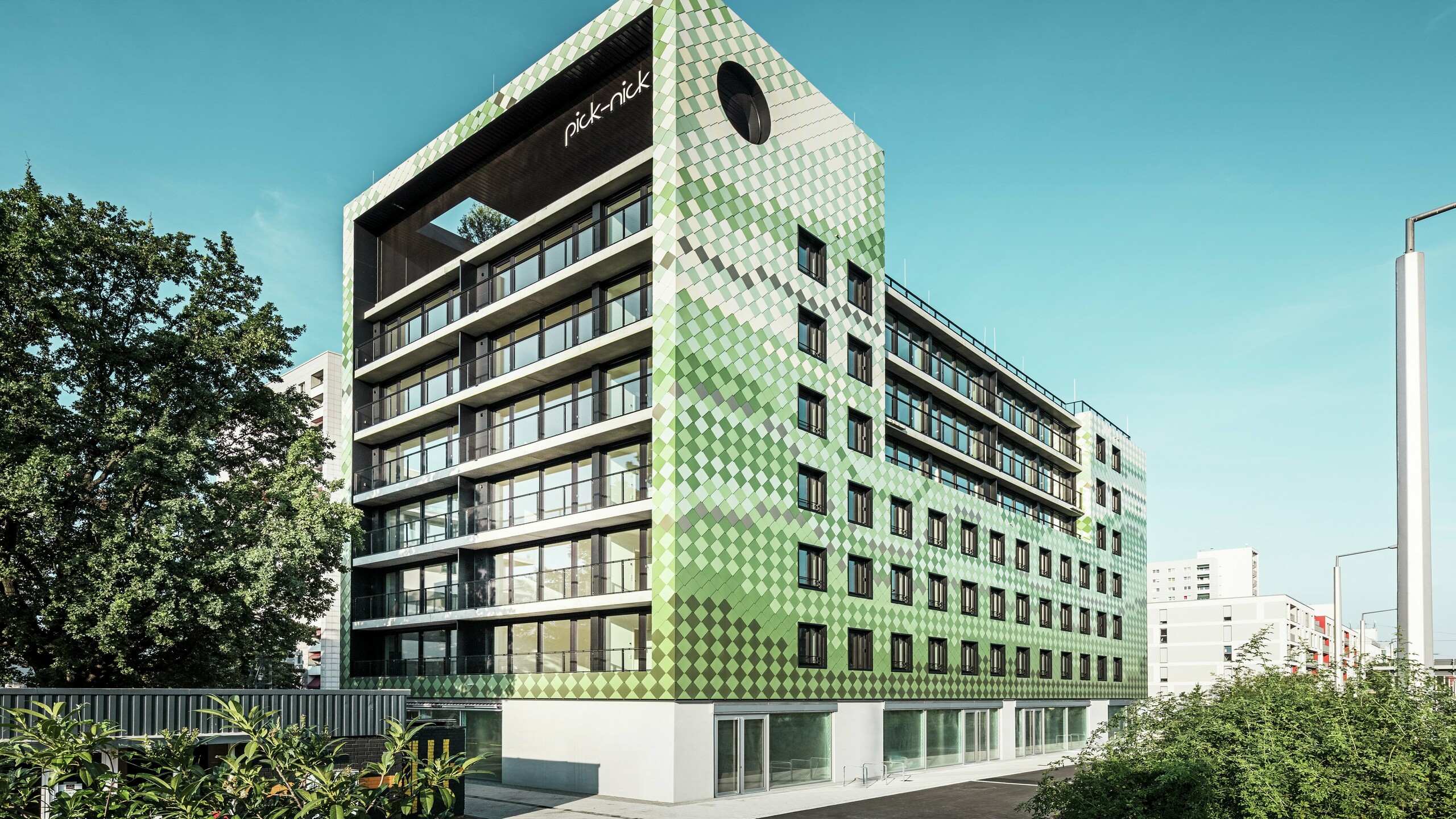 The ‘Pick-Nick’ residential and commercial building in Dresden boasts a modern PREFA aluminium façade made of rhomboid panels in various shades of green and beige. The striking building envelope combines clear lines with a dynamic play of colours and is complemented by generous glass surfaces. The ‘Pick-Nick’ lettering on the upper edge of the building and the green roof terrace add architectural highlights.