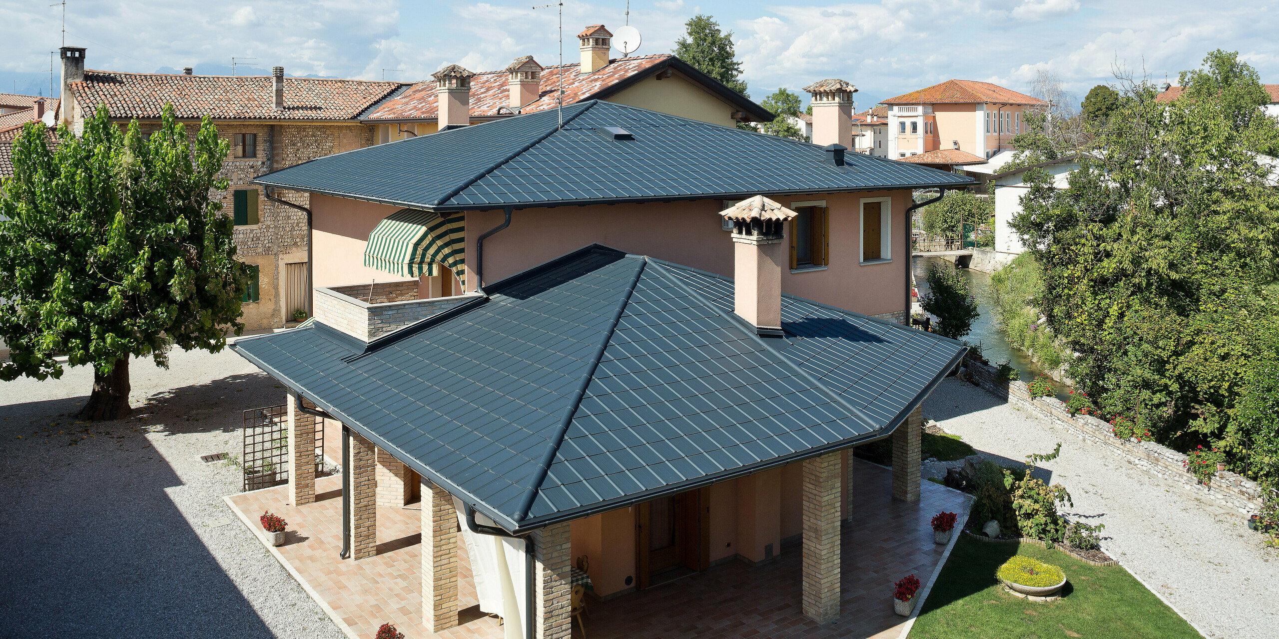 Elegant roof design in San Lorenzo di Arzene: Villa Beroia relies on PREFA P.10 roof tiles in anthracite. The building thus combines Italian tradition with a modern aluminium roof. The surrounding area features gravel paths, trees and shrubs, as well as a small river that flows directly past the house.
