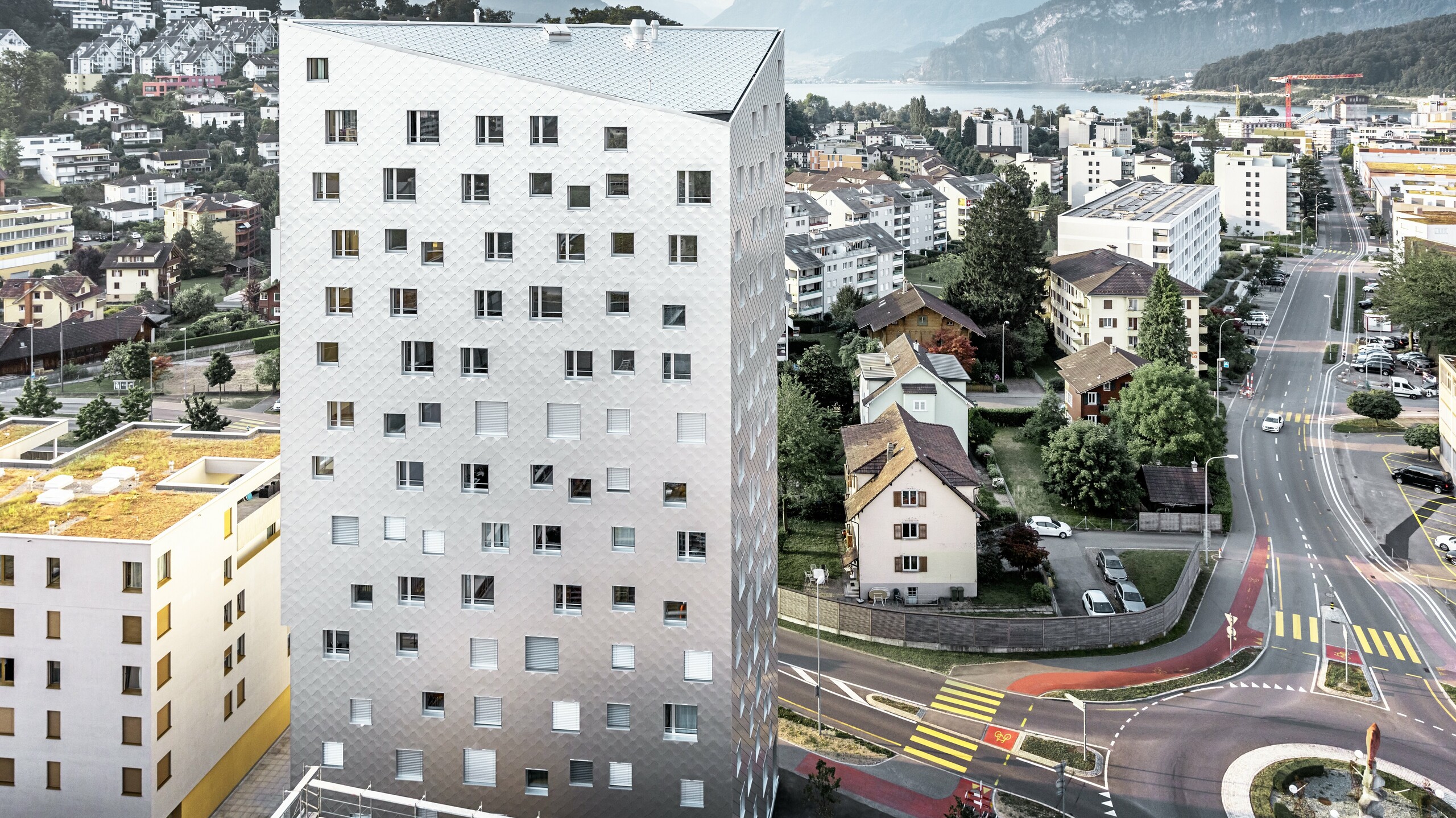 Das Bild zeigt das Solitaire-Hochhaus in Horw, einem Vorort von Luzern, das von der Schweizer Architektin Tilla Theus entworfen wurde. Das Gebäude hat unregelmäßig angeordnete Fenster, die als 'tanzende Fenster' bekannt sind, und ist mit einer Fassade aus eloxierten PREFA Aluminiumrauten verkleidet, die die Umgebung und die Wolken reflektieren. Im Hintergrund sind weitere Gebäude der Stadt, Straßen und die malerische Schweizer Berglandschaft zu sehen.