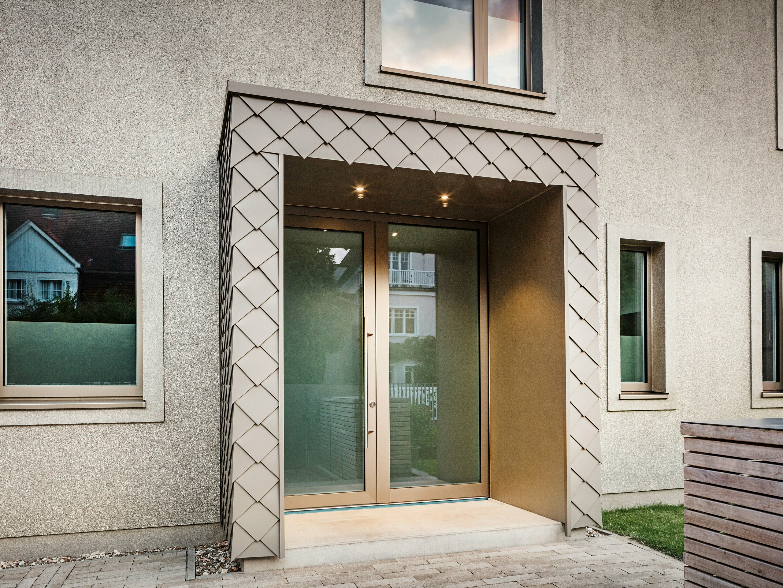 Close-up of the entrance area of a modern detached house with bronze-coloured aluminium cladding from PREFA. The entrance is covered and clad with elegant aluminium rhomboid tiles, giving the area a modern and stylish look. There are large windows to the left and right of the entrance door, creating a bright and inviting atmosphere.