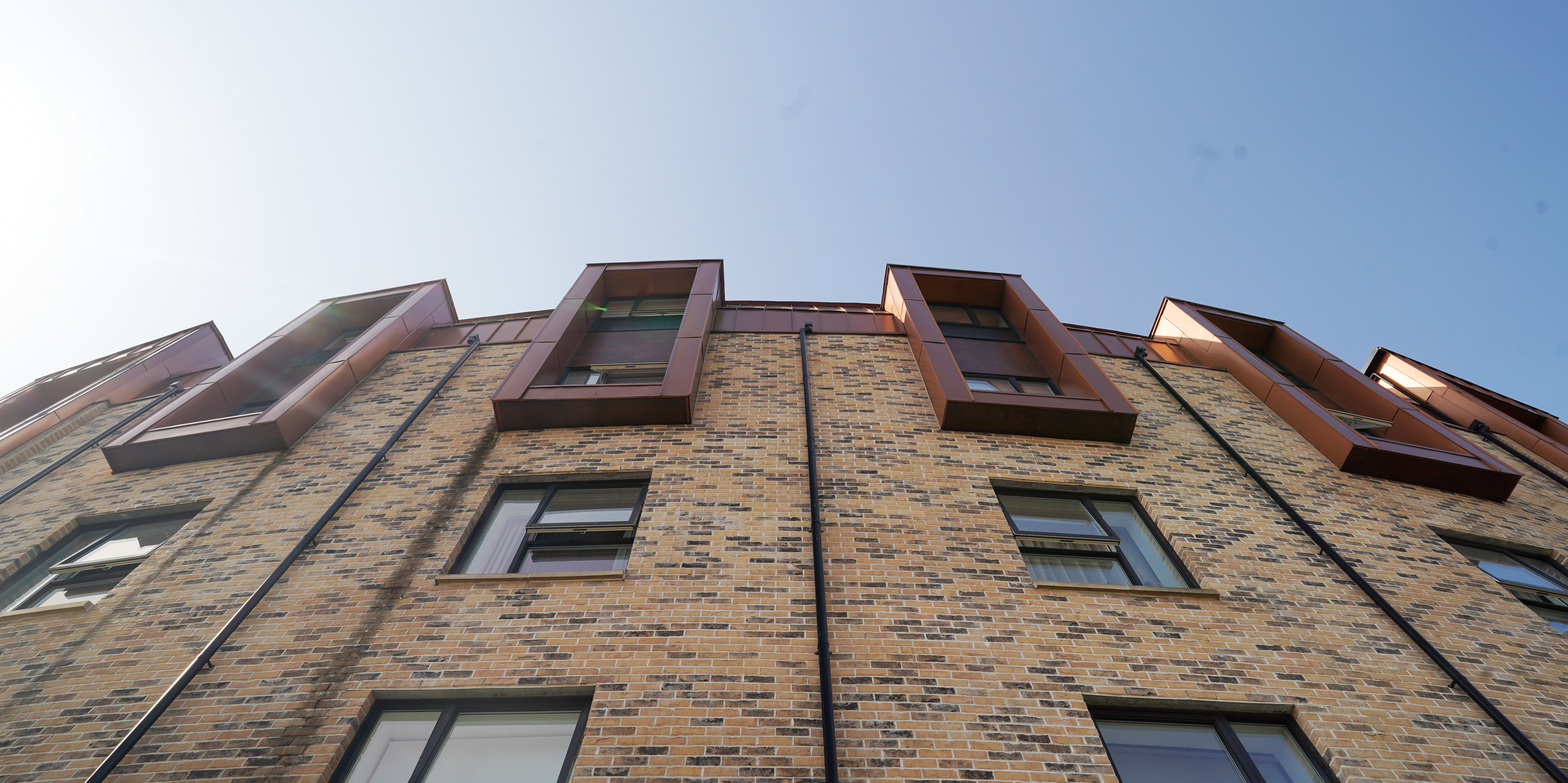 An upward view of the façade of a residential development on Nethan Street in Glasgow, which features the use of FALZONAL in New Copper, a durable and robust PREFA aluminium product. The vertical alignment of the windows, framed with the copper-coloured aluminium, breaks up the horizontal articulation of the brick wall and gives the building a dynamic and modern look. The quality and colouring of the FALZONAL blends seamlessly into the urban architecture while offering a durable and aesthetic solution for modern residential complexes. Black downpipes for roof drainage have been placed between the windows, emphasising the curvature of the building from this perspective.