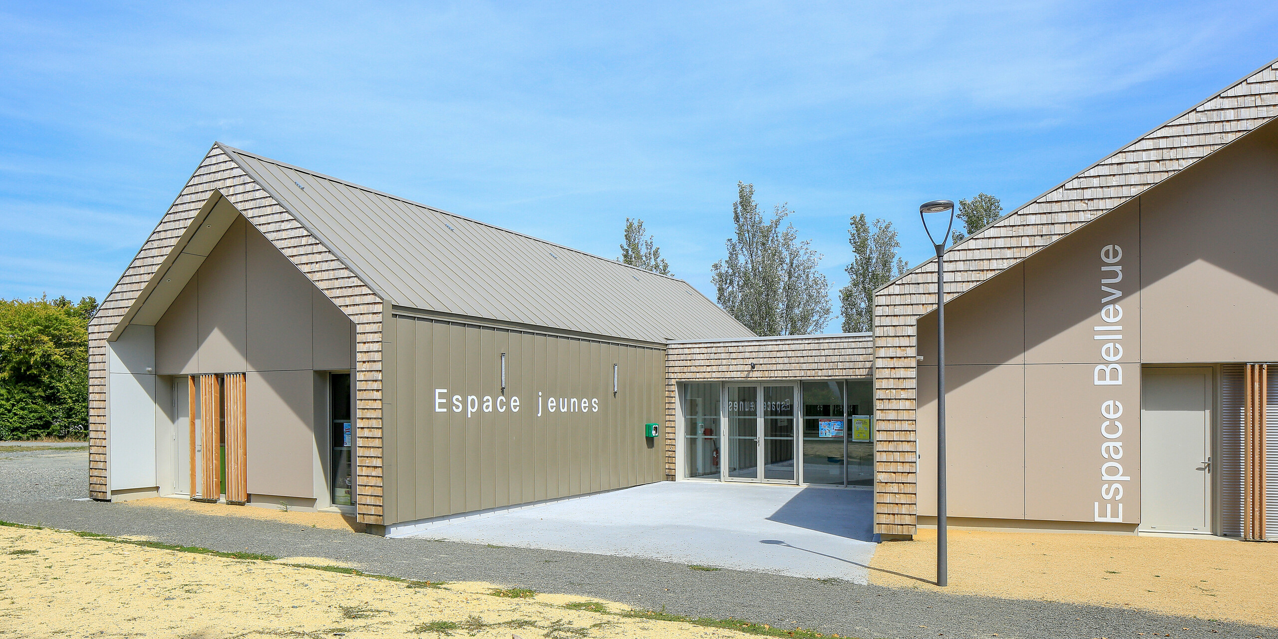 Salles Espace Bellevue in Mûrs-Erigné, Frankreich, mit den Schriftzügen 'Espace Jeunes' und 'Espace Bellevue' vor dem Veranstaltungssaal. Das PREFALZ Dach- und Fassadensystem in Bronze schmückt große Teile des modernen Jugendzentrums. Die witterungsbeständige Aluminium-Stehfalzverkleidung von PREFA bietet eine langlebige und moderne Lösung für Bauvorhaben. Die Kombination aus Aluminiumverkleidung und natürlicher Holzoptik fügt sich harmonisch in die Umgebung ein.