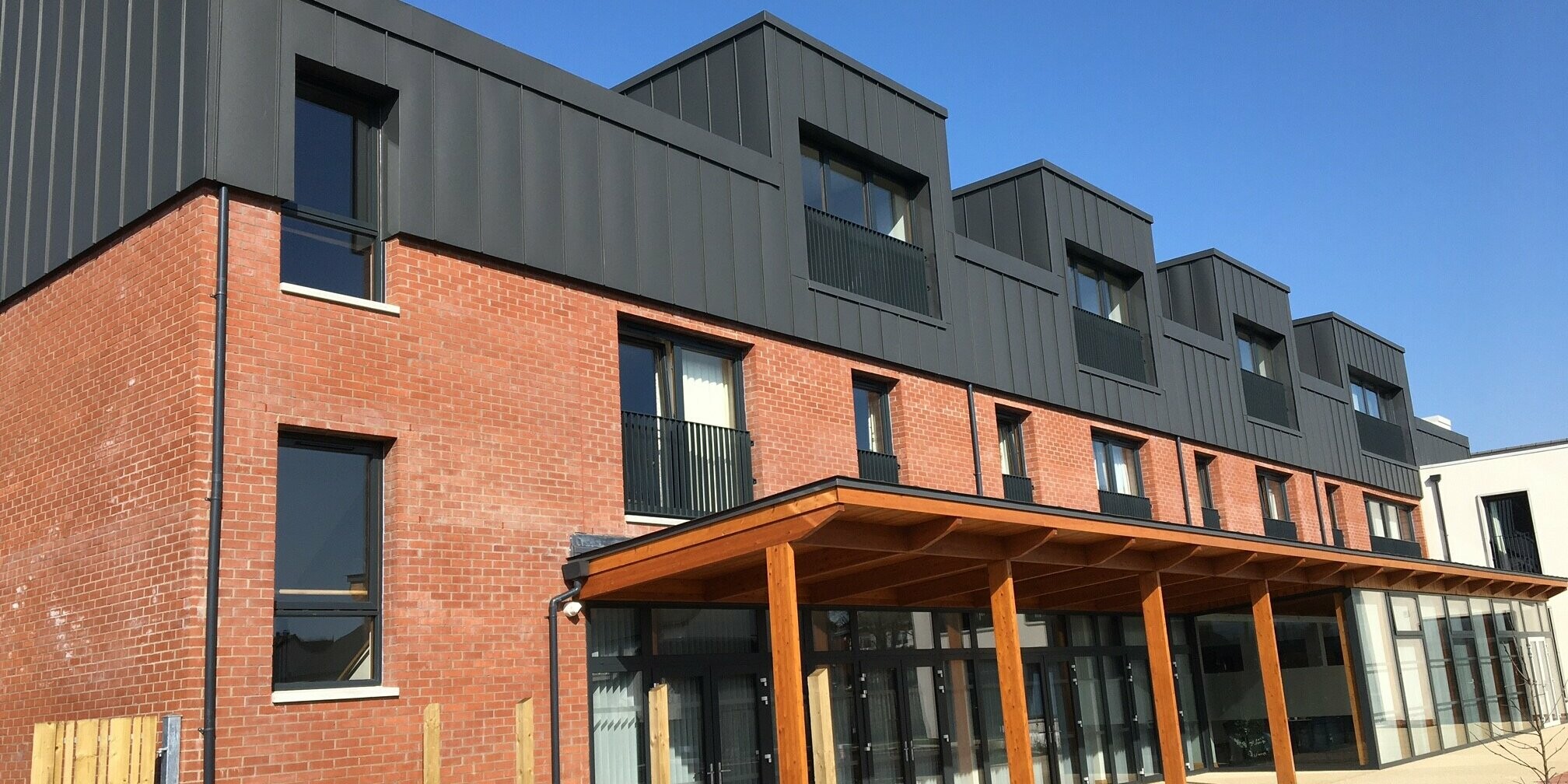 View from the garden of Harberton Hall in Belfast, Northern Ireland. The standing seam roof made of dark grey PREFALZ aluminium matches the brick façade. A green outdoor area and flat roof dormers enhance the harmonious combination of modern architecture and traditional style in this sheltered housing complex.