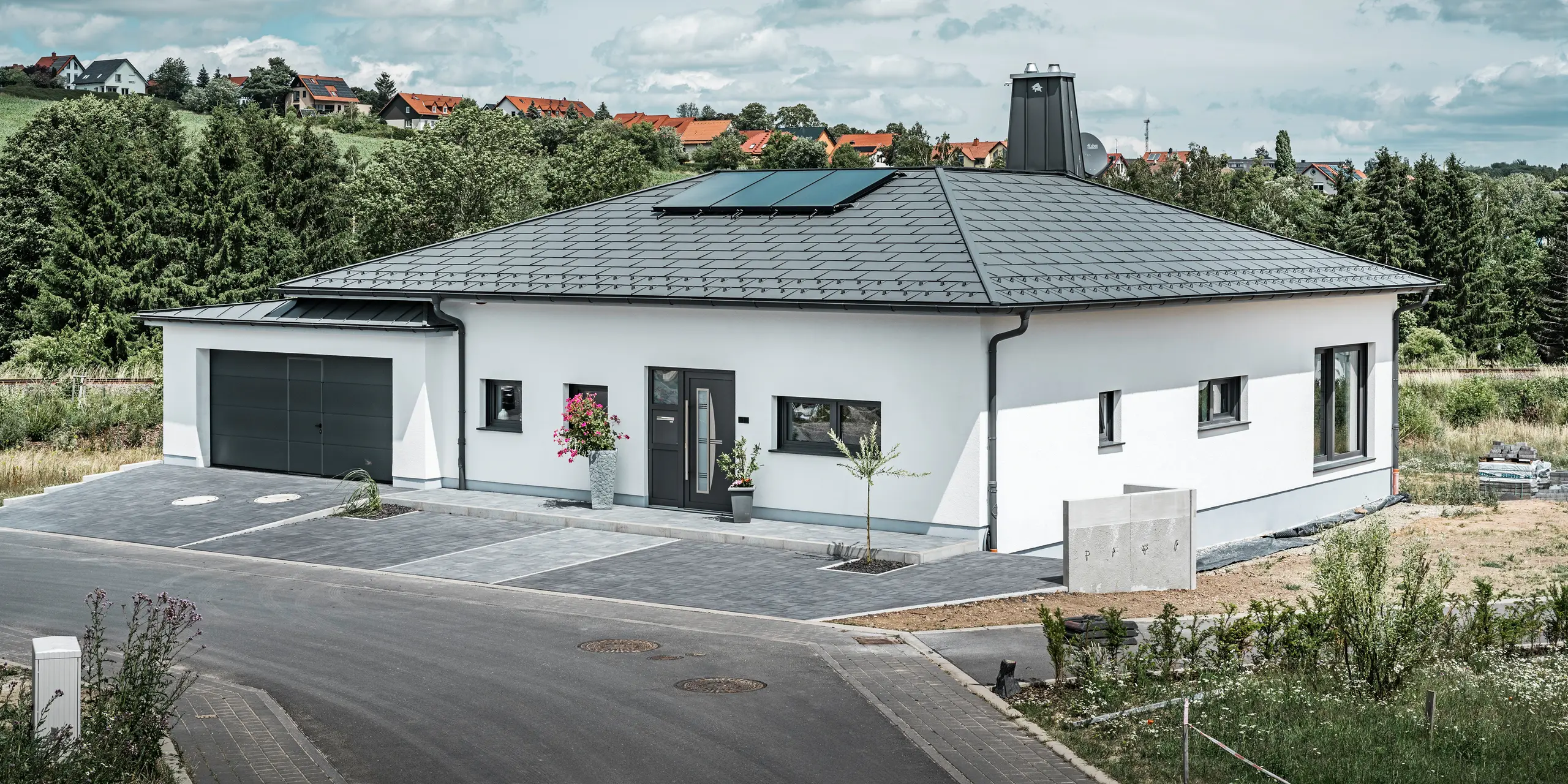 Elegant detached house in Ilmenau, Thuringia, with a high-quality PREFA roof in P.10 dark grey, fitted with robust R.16 roof tiles. The garage harmonises in colour and style thanks to the use of PREFALZ. The eye-catching features of the property are the functionally designed atrium and the chimney decorated with the PREFA bull, which underline PREFA's modern design and innovative roof technology. The solar panels fit seamlessly into the sustainable concept of the house. The well-thought-out overall concept of the bungalow is rounded off by the PREFA roof drainage system, which is also in dark grey.