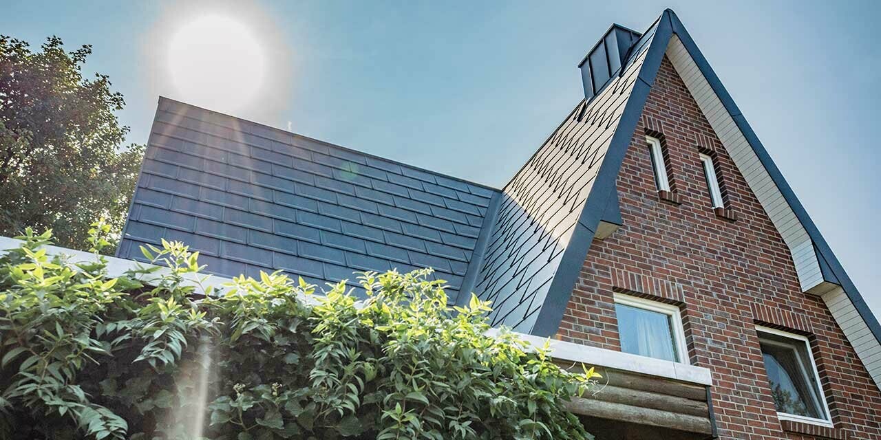 Gabled roof clad in the plain PREFA R.16 aluminium roof tile in anthracite. The house has a rustic brick façade – the photo is taken as the sun rises behind the house.