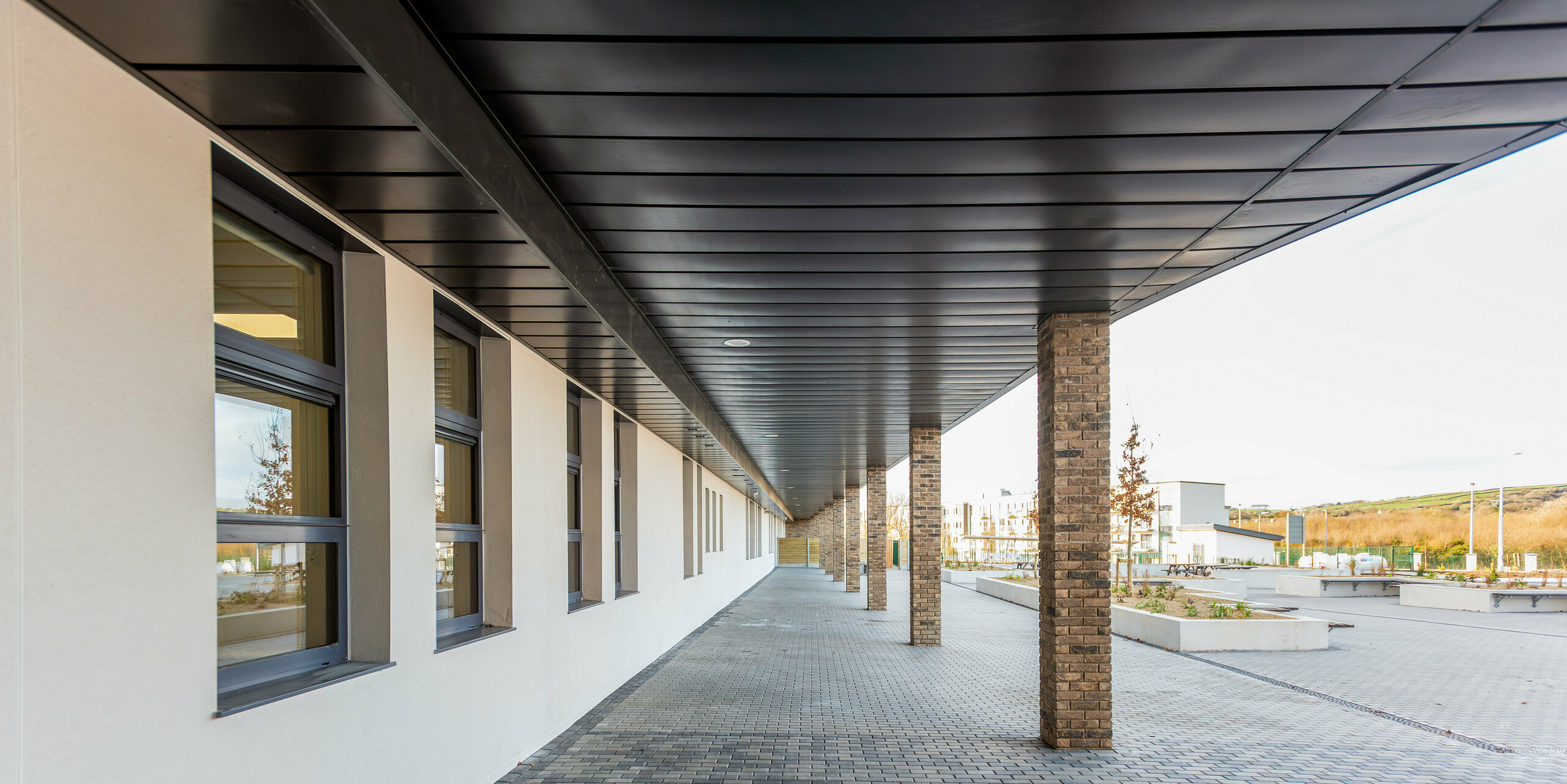 Porch with brick columns at the entrance of the Scoil Mhuire Naofa in Carrigtwohill, Ireland. The high-quality PREFALZ cladding in P.10 light grey also ensures an elegant appearance under the roof, ideally combined with the robust brick columns and the timeless architecture.