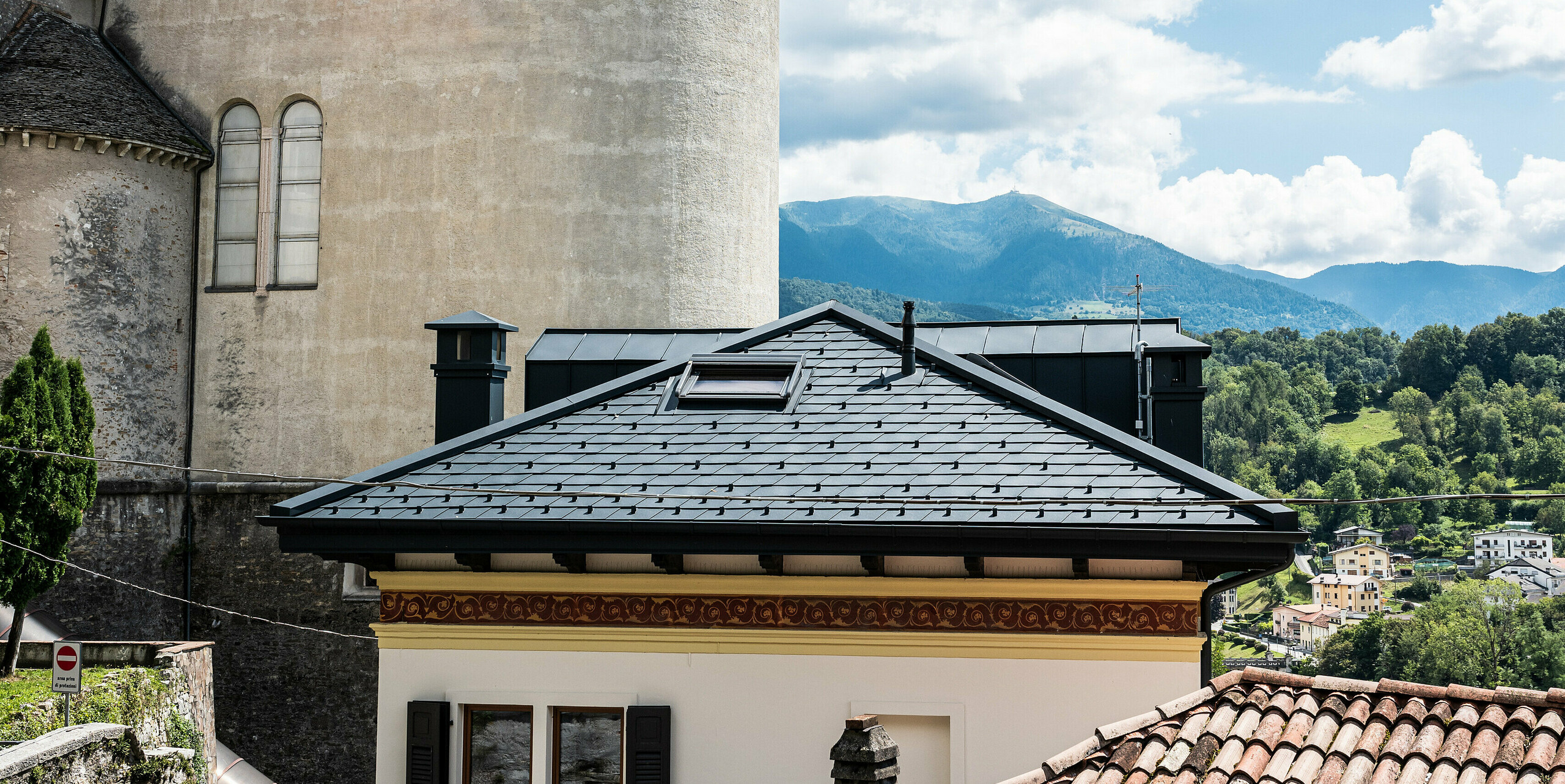 Historische und moderne Architektur vereint in Belluno, Italien, mit einem sorgfältig modernisierten Wohngebäude, dessen Dach mit anthrazitfarbenen PREFA Aluminiumdachschindeln versehen ist. Das Gebäude steht im Einklang mit der umgebenden traditionellen Struktur und dem grünen, hügeligen Hintergrund, während die klare Linienführung und die Kaminverkleidungen das zeitgenössische Design und die innovative Nutzung von langlebigen Materialien hervorheben.