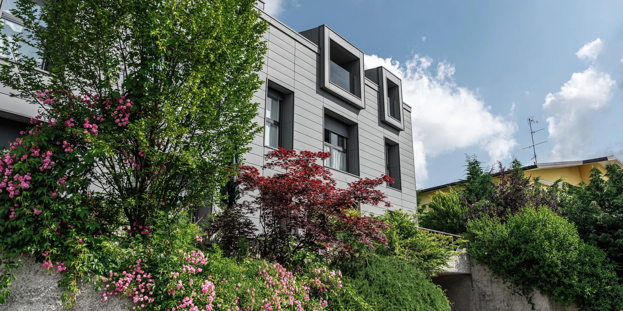 Modern residential complex in Faloppio, Italy, with a stylish façade clad in PREFA siding. The cantilevered windows provide a unique architectural feature, while the surrounding plants and flowers emphasise the natural beauty of the property. The durable aluminium cladding from PREFA provides an aesthetic and functional solution in all weather conditions.