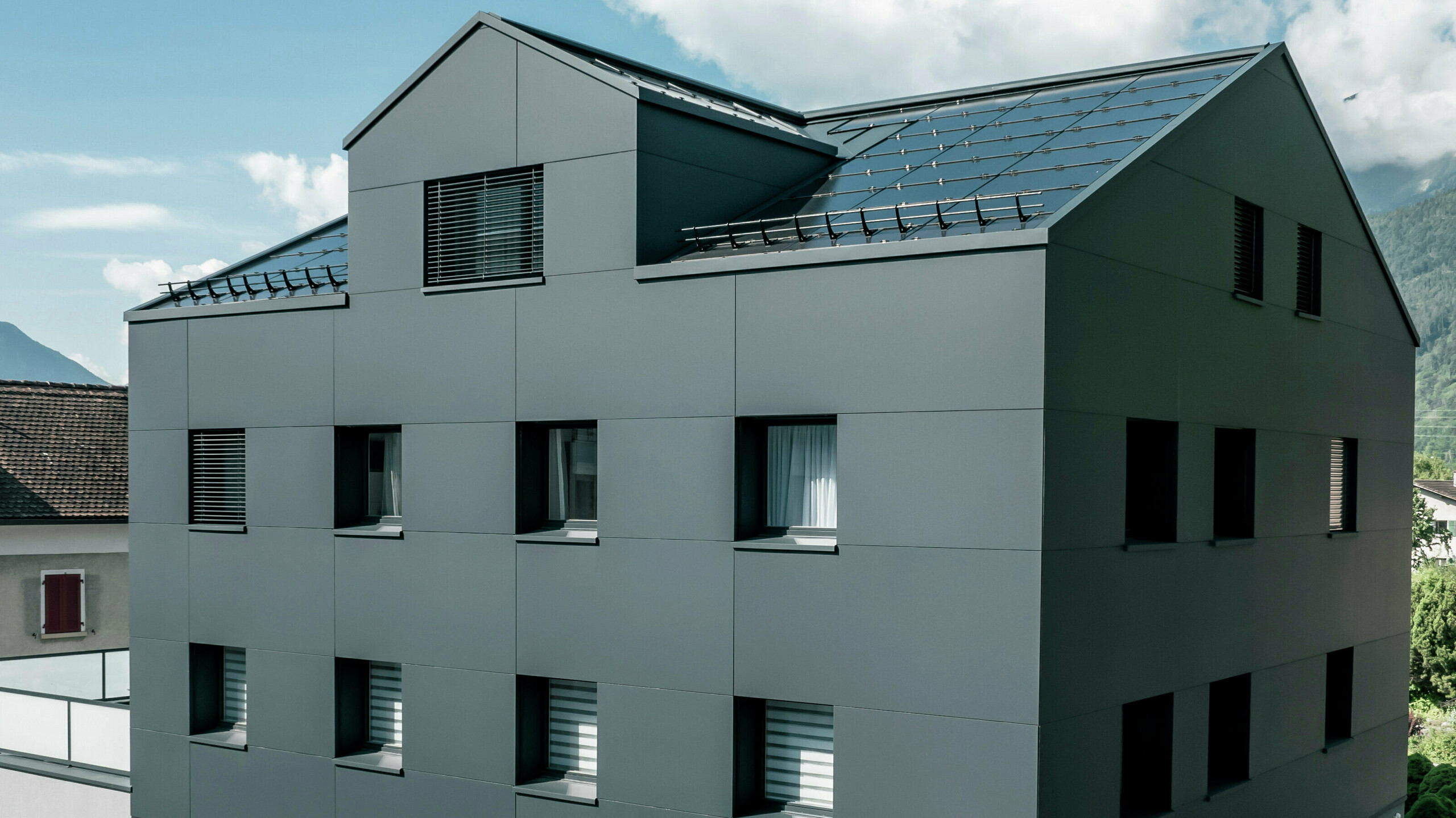 Modernisiertes Wohn- und Geschäftsgebäude nach der Fassadensanierung mit PREFABOND Aluminium Verbundplatten in Anthrazit. Die klare, elegante Linienführung der neuen Fassade verleiht dem Gebäude ein modernes Erscheinungsbild. Im Erdgeschoss befindet sich das Architekturbüro Voser. Die Umgebung mit Bäumen und Bergen rundet die harmonische Optik ab.